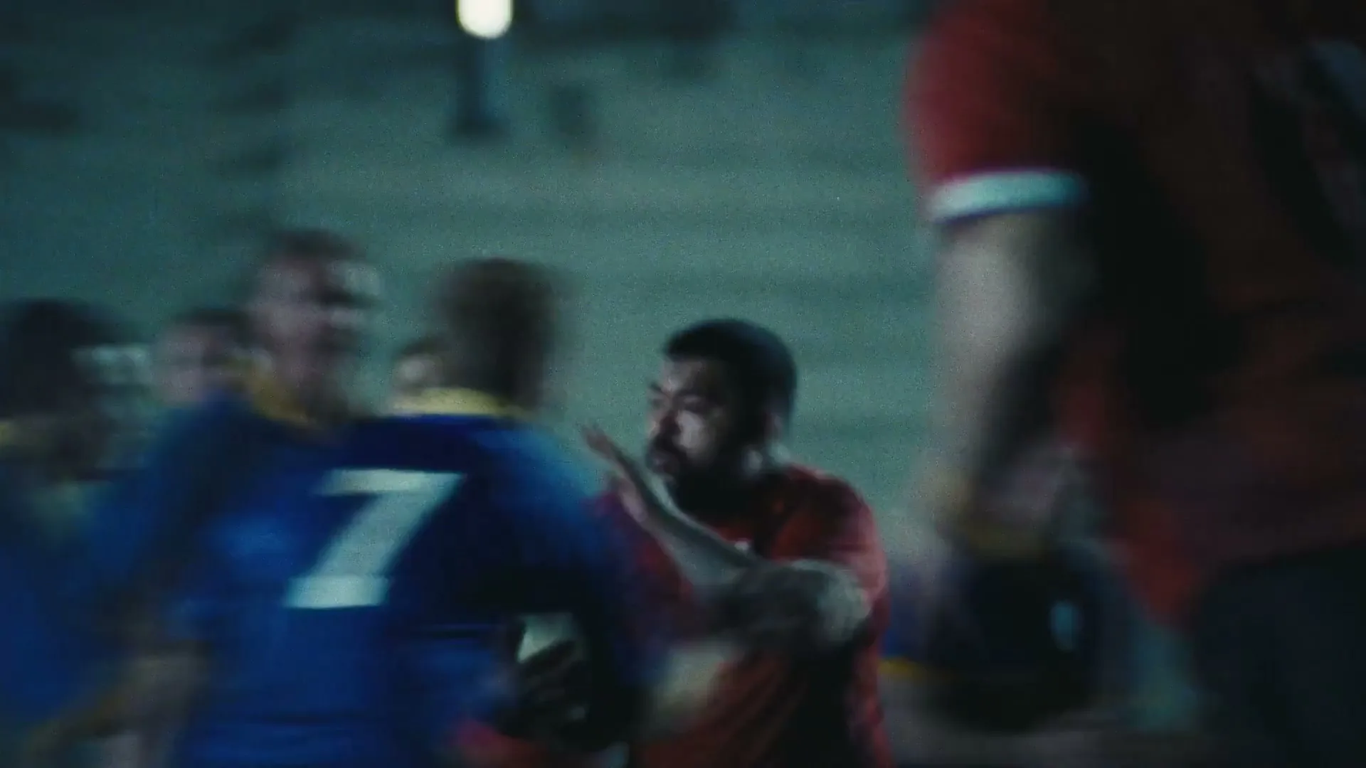A group of male rugby players in blue and red jerseys are playing on a field at night, with one player in red running with the ball.