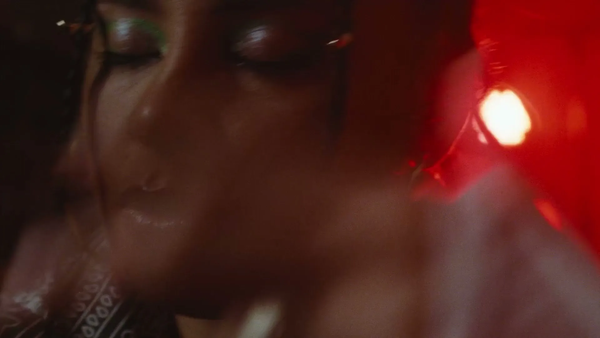 A young woman with braids and green eyeshadow closes her eyes as a hand moves across her face in a dimly lit room.
