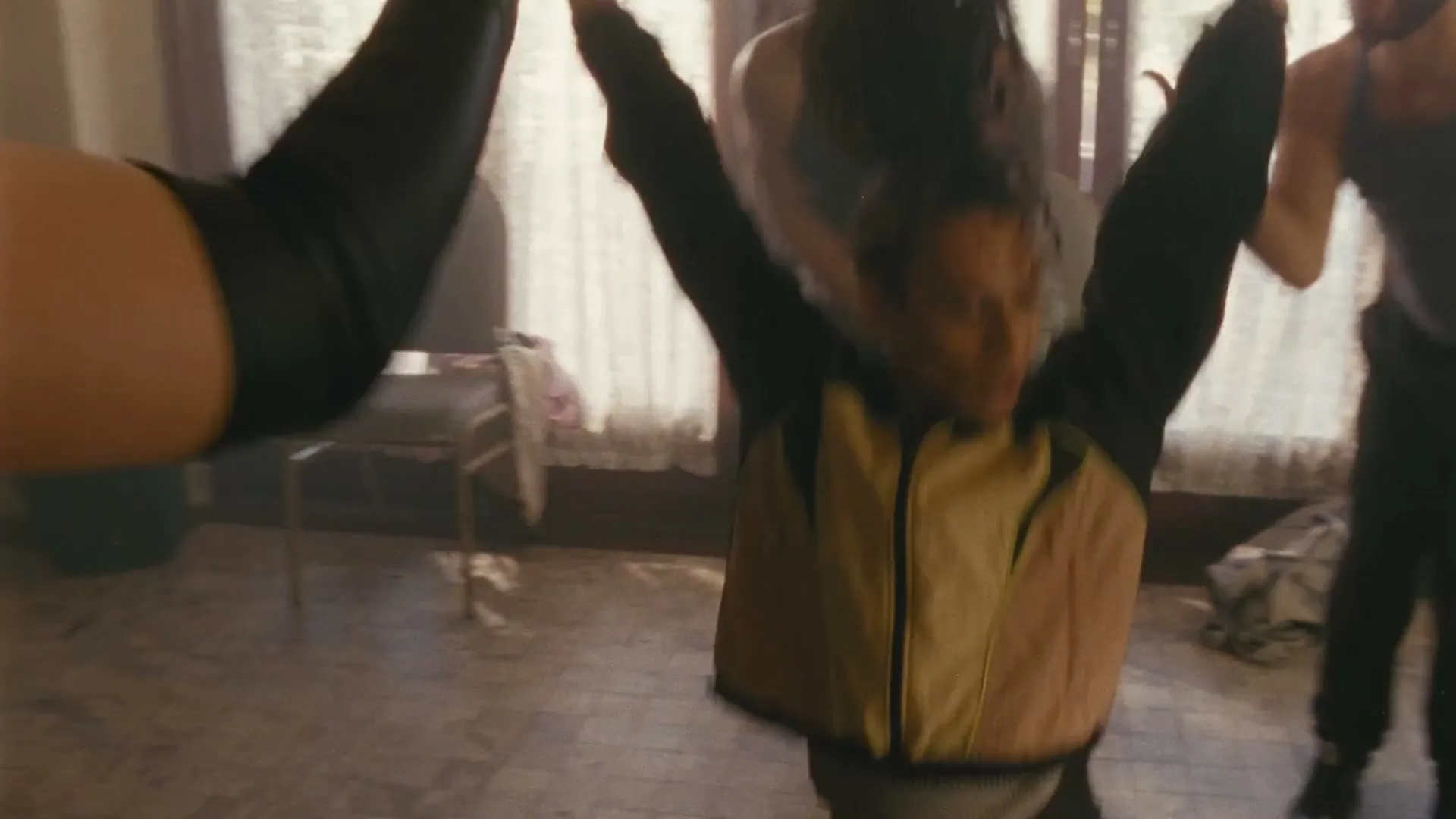 A person with curly hair in a black and tan jacket dances with arms raised, surrounded by other people in a sunlit room.