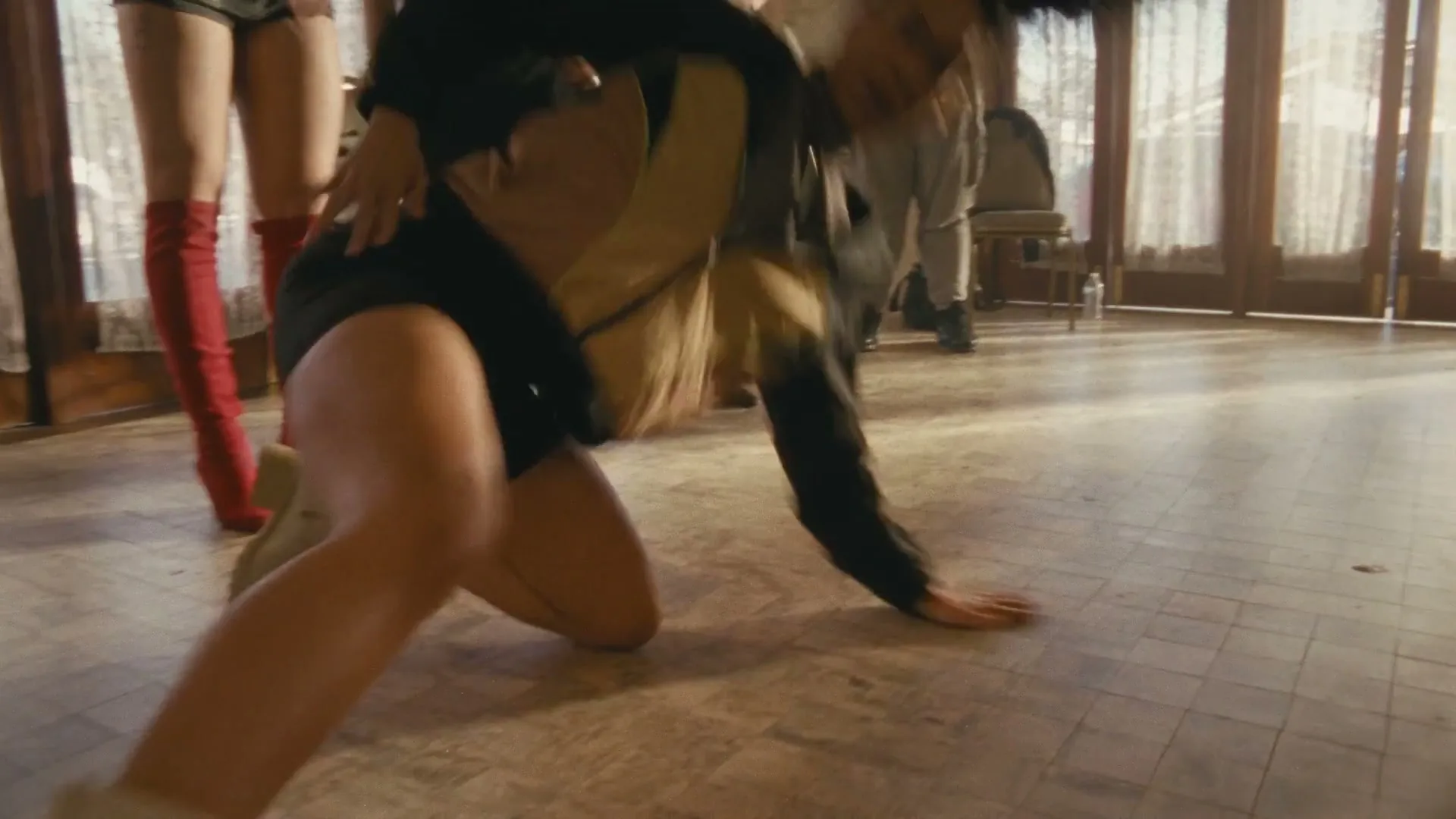 A diverse group of people in a dance studio watch a woman in a black and yellow outfit fall to the floor.