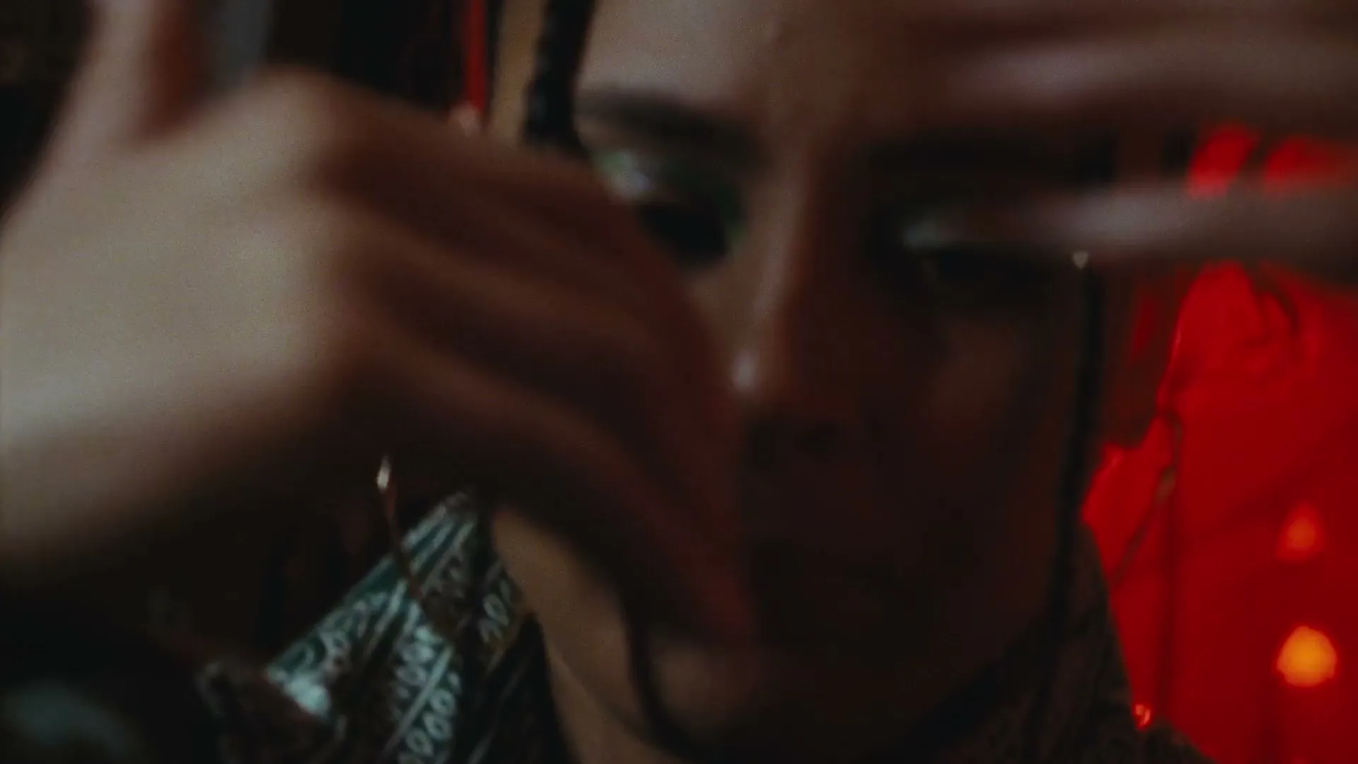 A person with green eye makeup and a patterned shirt moves their hands in front of their face, with red lights in the background.
