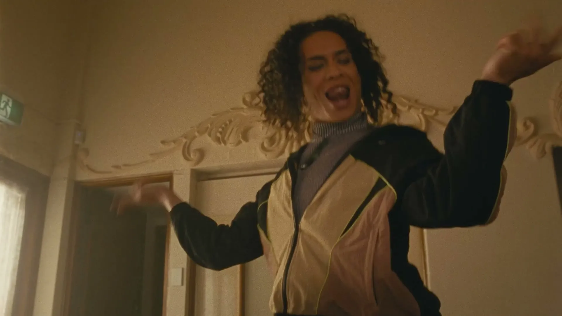 A person with curly hair and a colorful jacket dances energetically in a room with ornate wall decorations.
