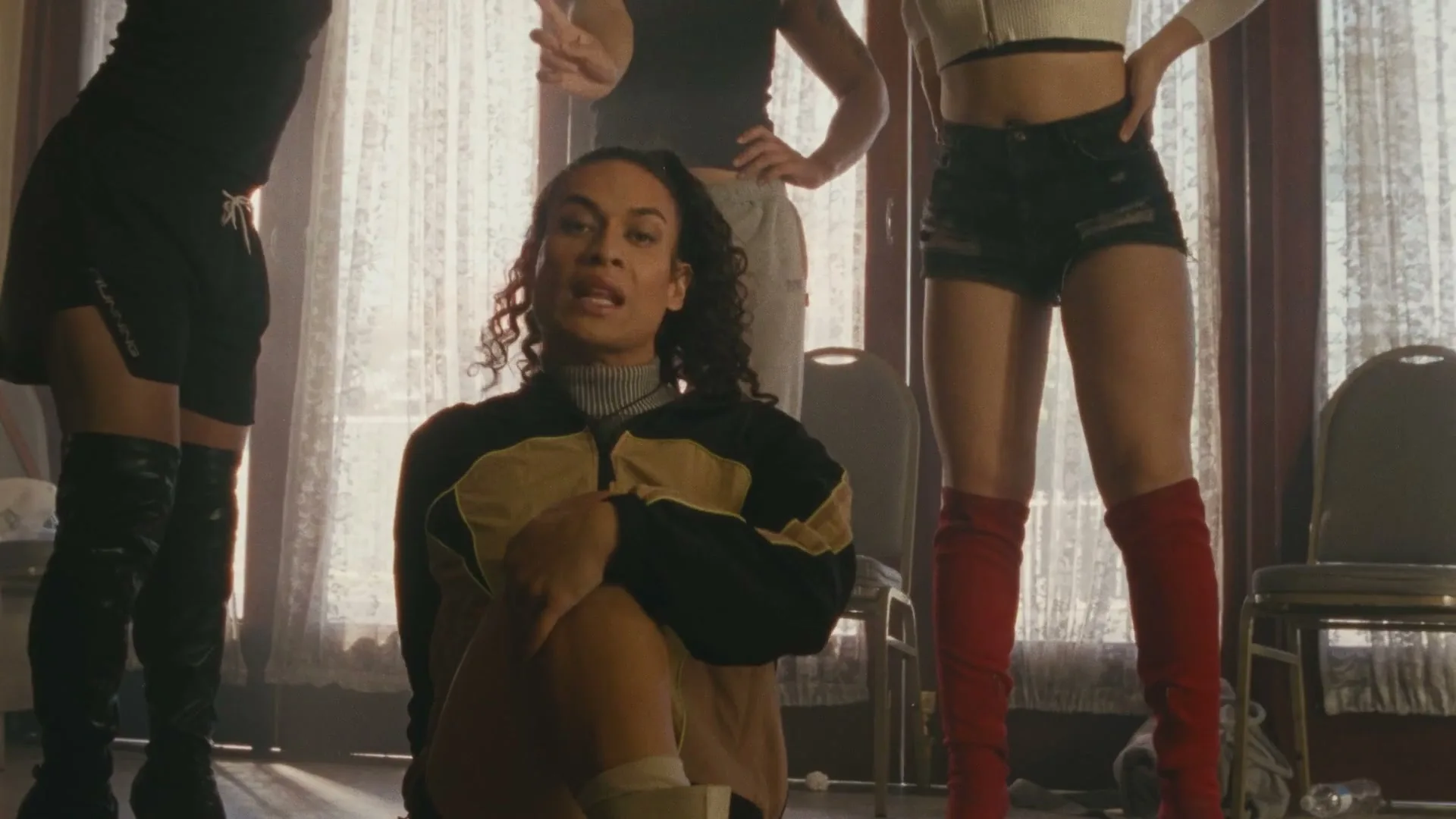 A person with curly hair sits on the floor, looking at the camera, surrounded by other people's legs in a room with sheer curtains.