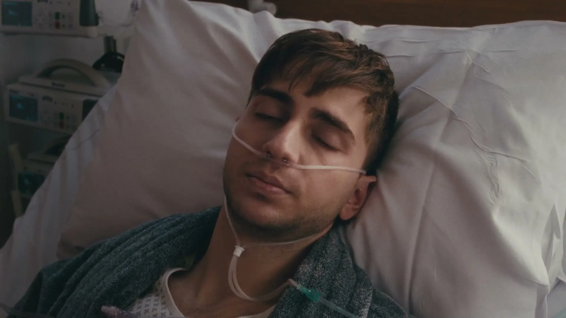 A young man with a nasal cannula lies in a hospital bed, looking directly at the camera with a somber expression.