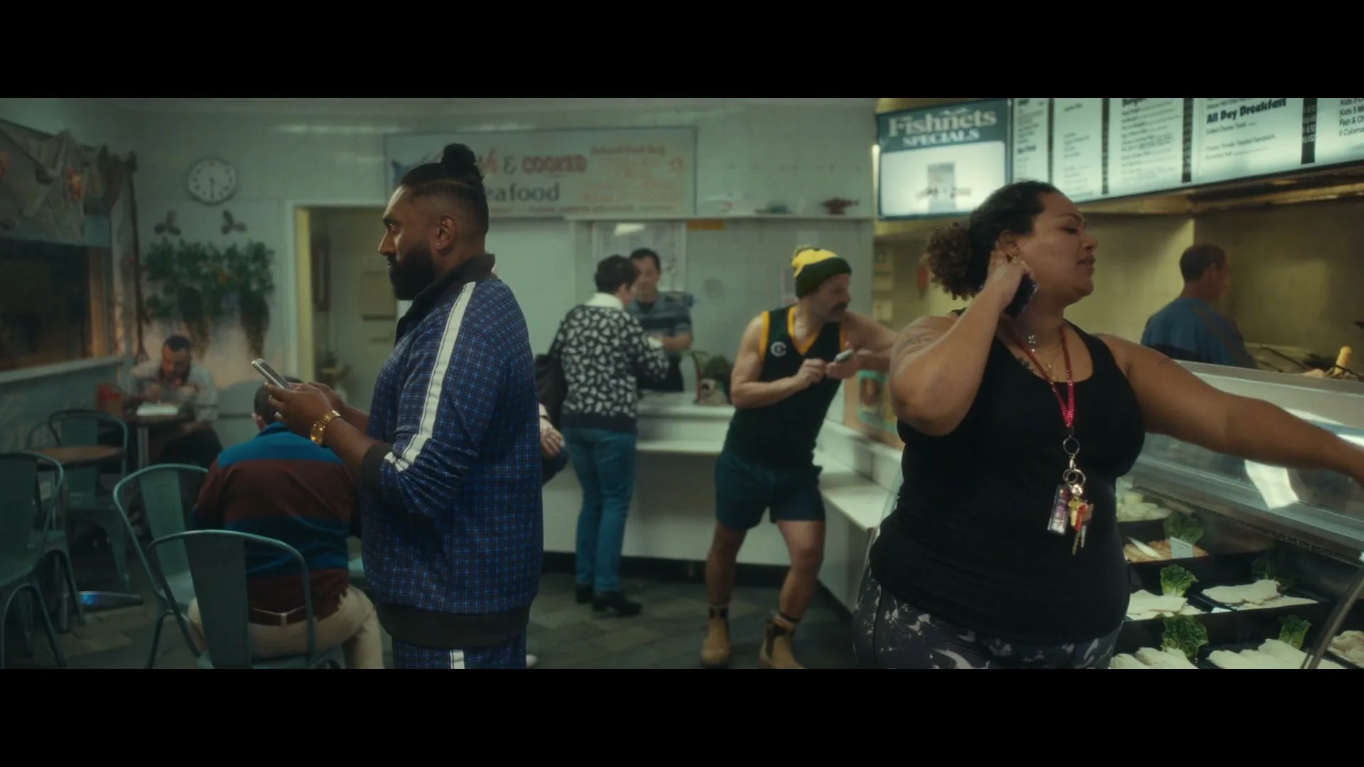 A diverse group of people are seen inside a fish and chips shop, with a man on the left looking at his phone and a woman on the right looking into a display case.