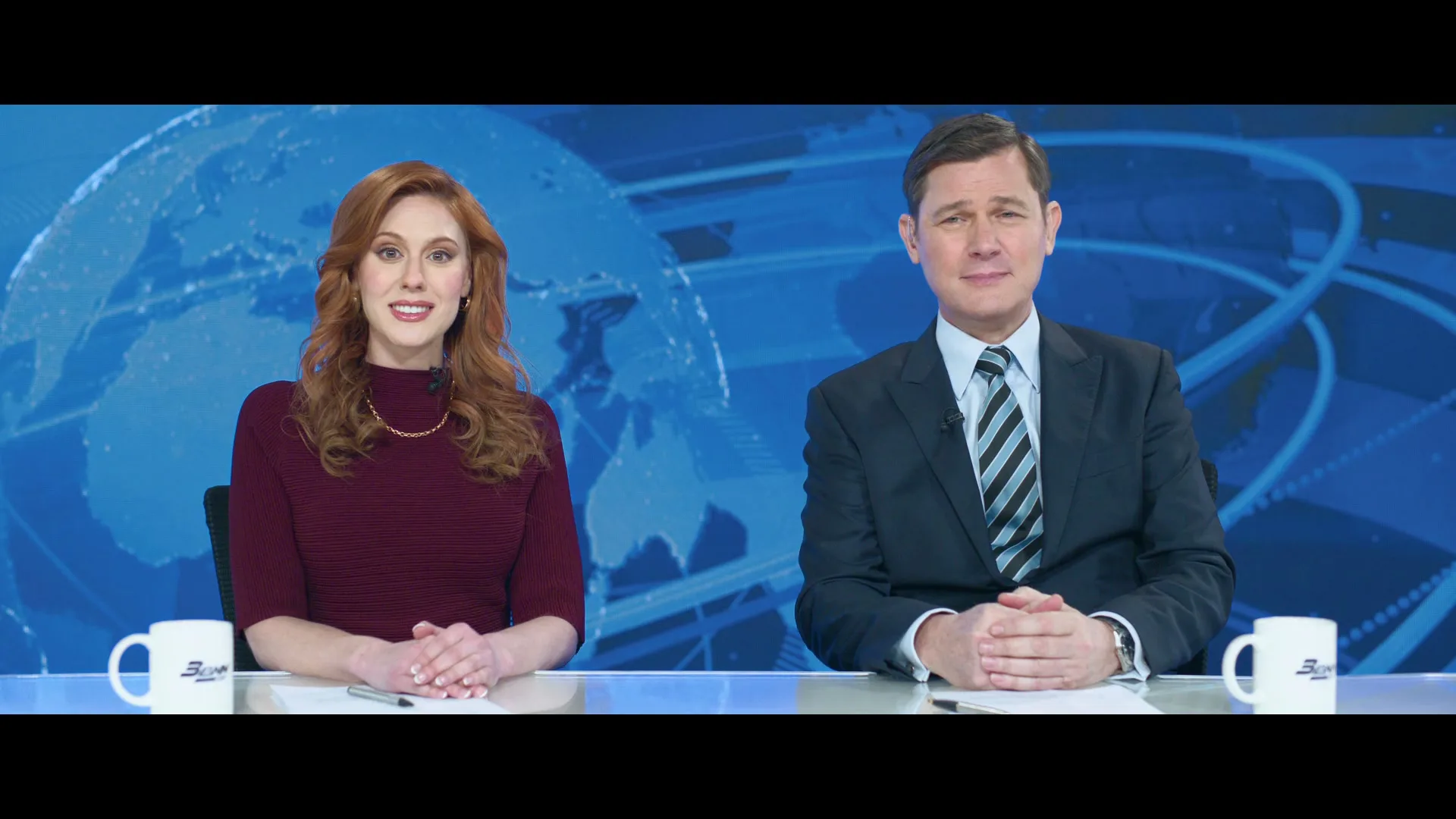 A female and male news anchor sit side-by-side at a desk in front of a blue screen with a globe graphic, looking at the camera.