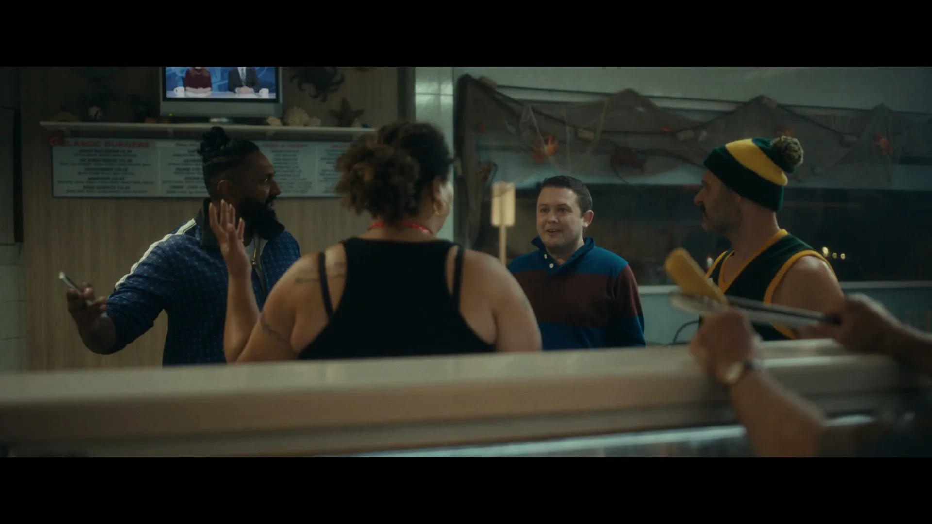 Four people, two men and two women, are talking in a fish and chips shop, with a TV playing in the background.