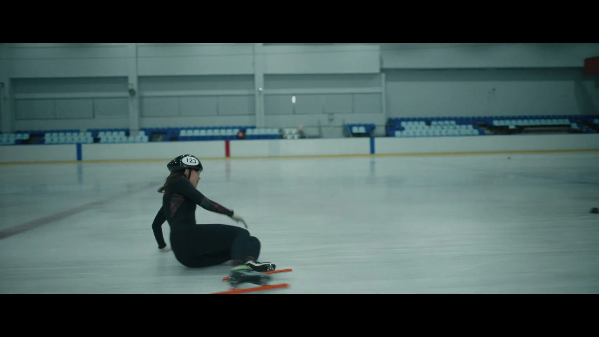 A female speed skater in a black suit and helmet falls on the ice rink during practice, captured in a medium shot.