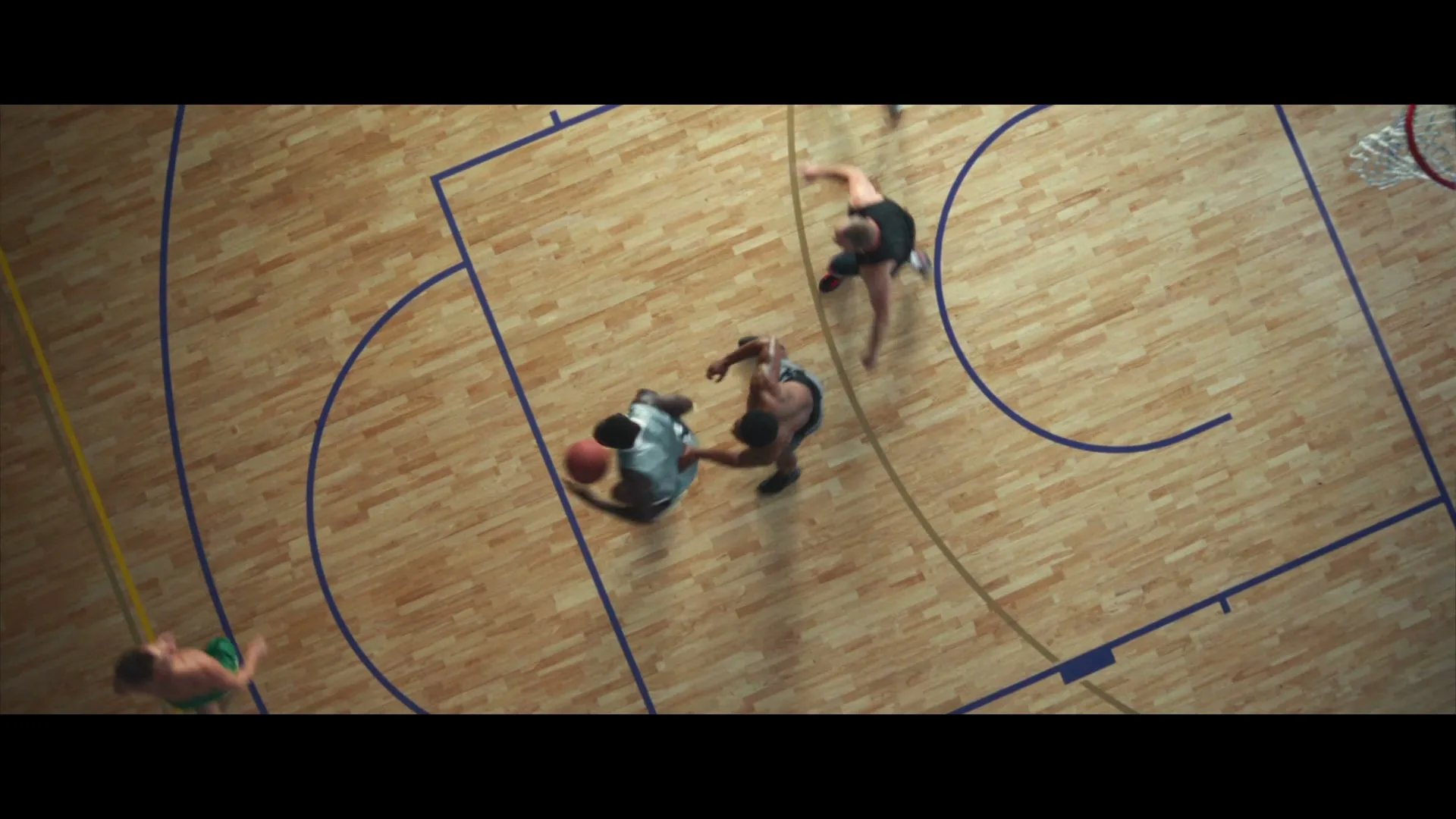 Four men play basketball on a wooden court, seen from an overhead perspective as one man dribbles the ball.