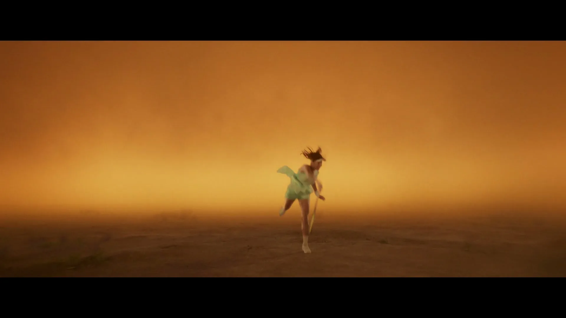 A female tennis player in a light green dress serves a tennis ball in a hazy, orange-lit, empty space, then transforms into a male cricket player.