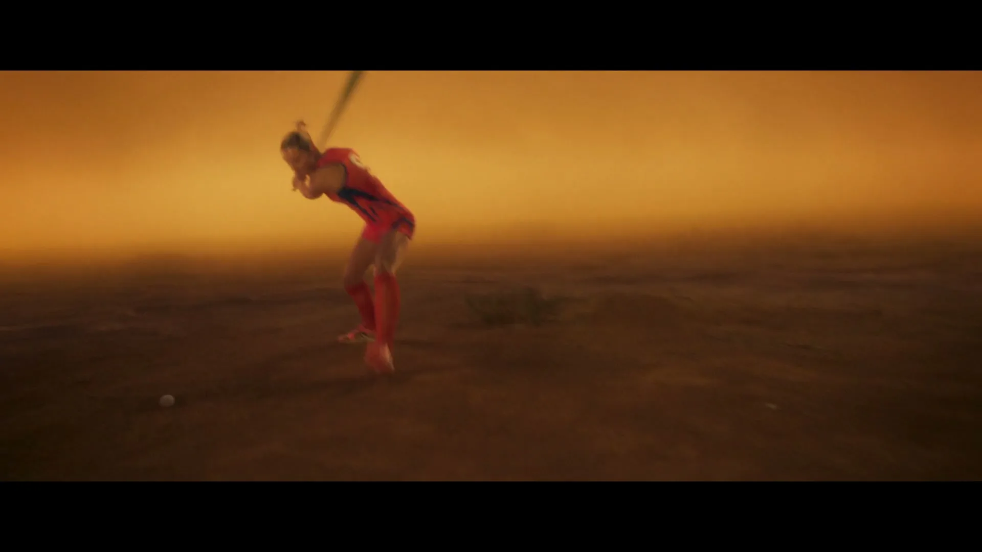 A female athlete in a red and blue uniform swings a cricket bat at a white ball on a dusty, barren field under an orange sky.