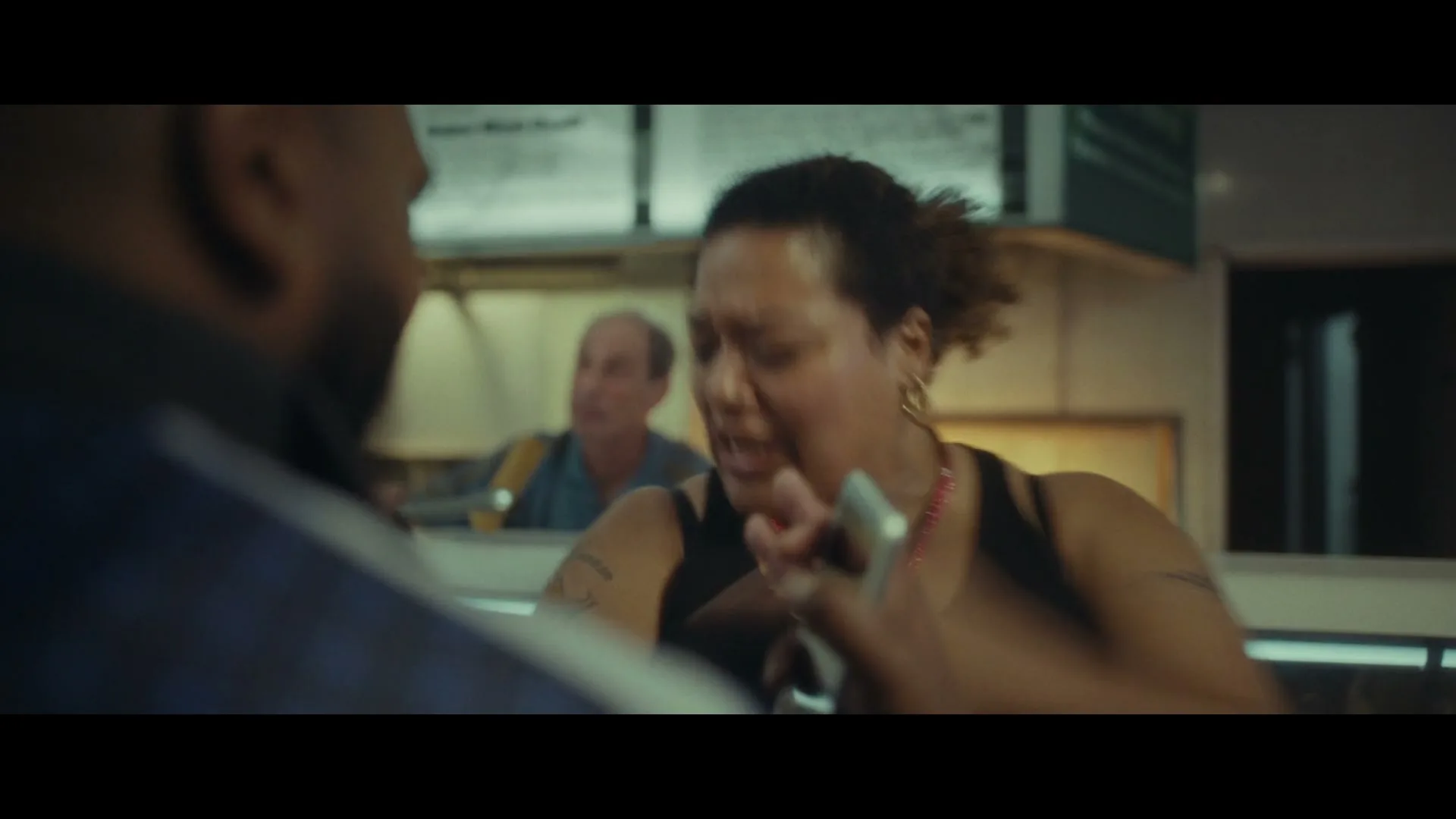 A woman in a black tank top and red beaded necklace argues with a man, while another man watches in the background at a fast food restaurant.