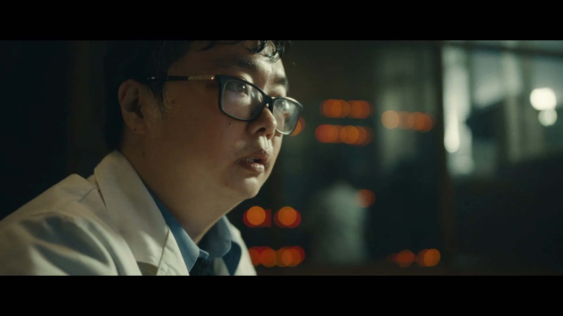 A man in a lab coat and glasses looks down and then up, with blurred orange lights in the background.
