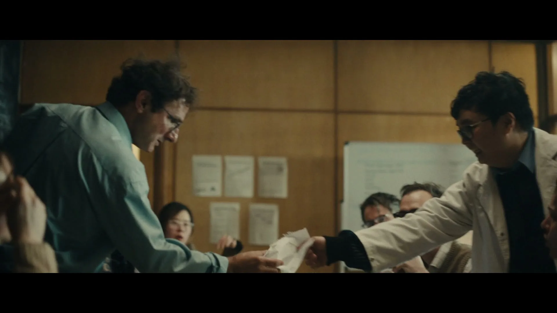 A group of scientists in a lab coats and glasses are in a heated discussion, passing papers around a table in a dimly lit room.