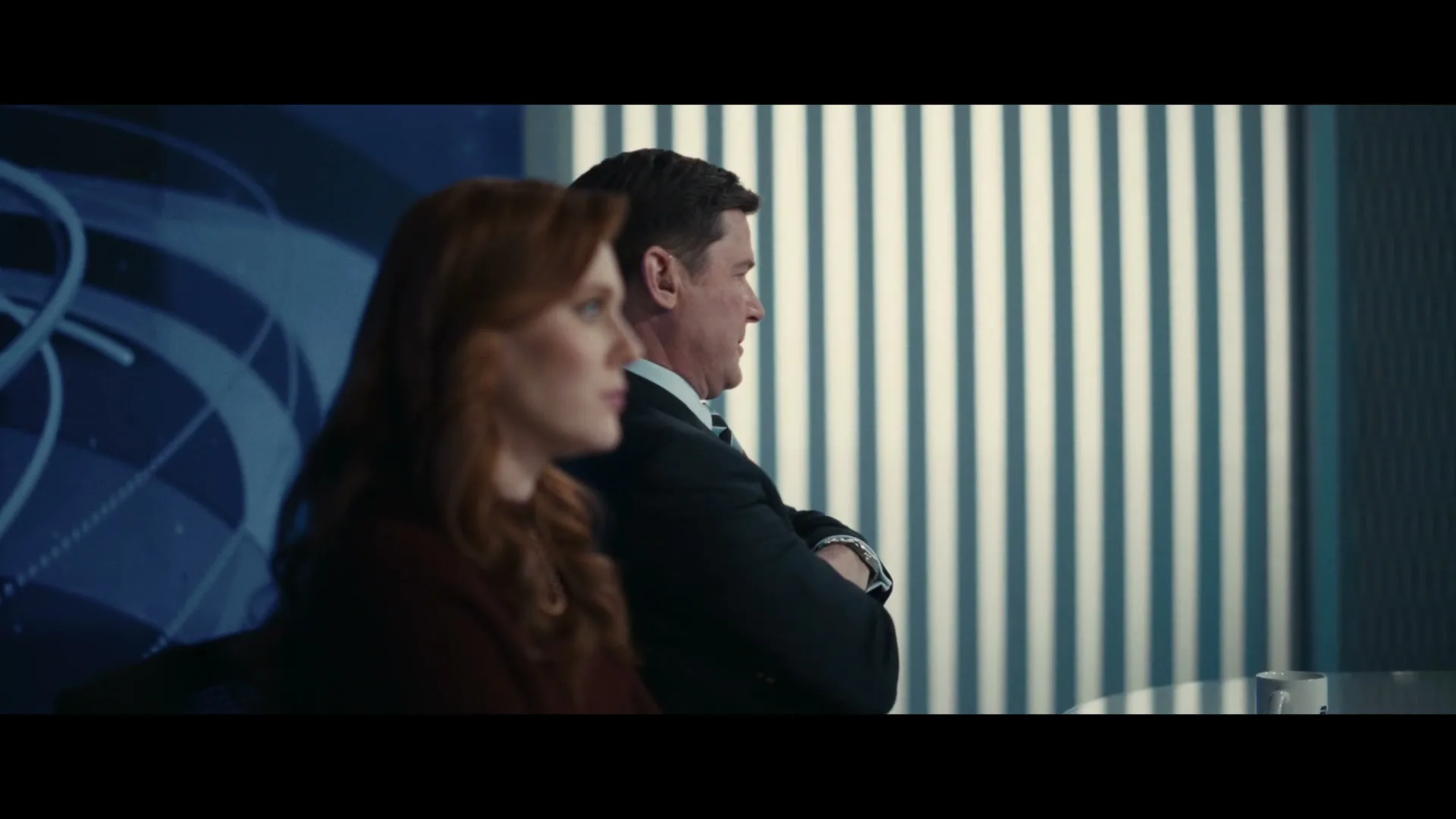 A man in a suit with crossed arms and a woman with red hair sit side-by-side, looking off-screen in a news studio.
