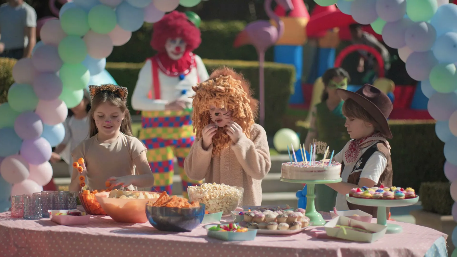 Three children in costumes stand around a table of snacks at an outdoor birthday party, with a clown and other partygoers in the background.