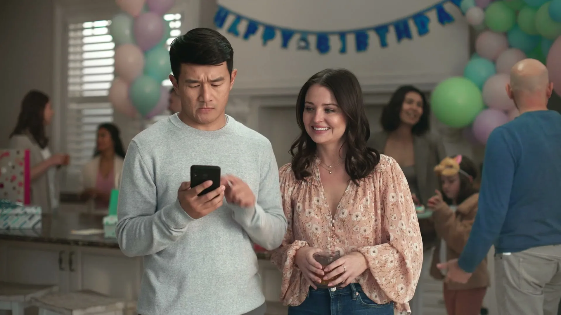 A man and woman stand together at a birthday party, with the man looking at his phone and the woman smiling, surrounded by other guests.
