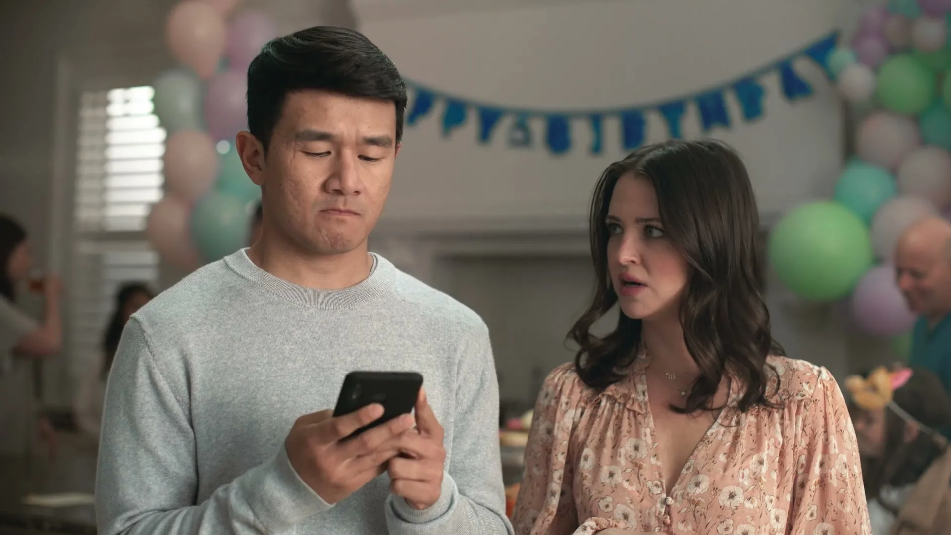 A man in a grey sweater looks at his phone while a woman in a floral dress looks at him with concern at a birthday party.
