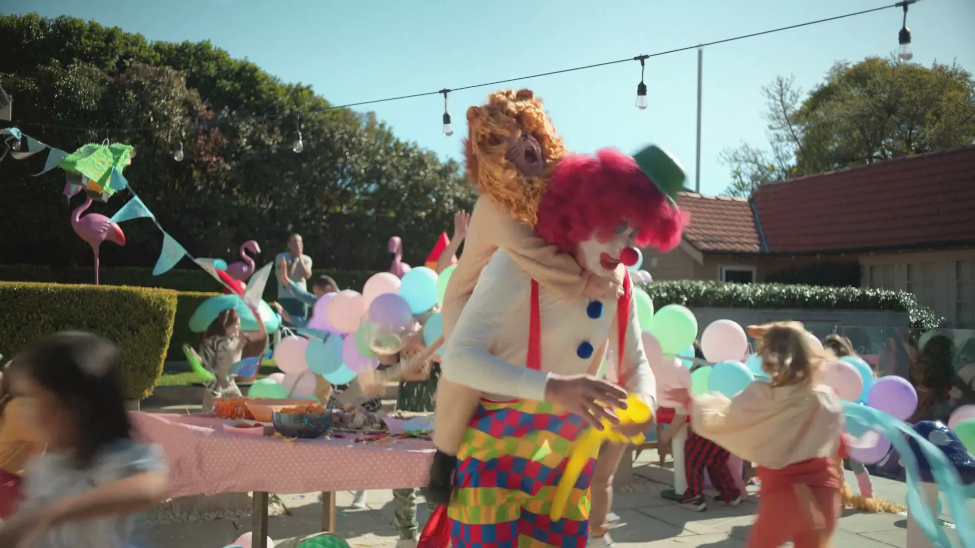 A clown in a colorful costume is giving a piggyback ride to a child in a lion costume at a chaotic outdoor birthday party.