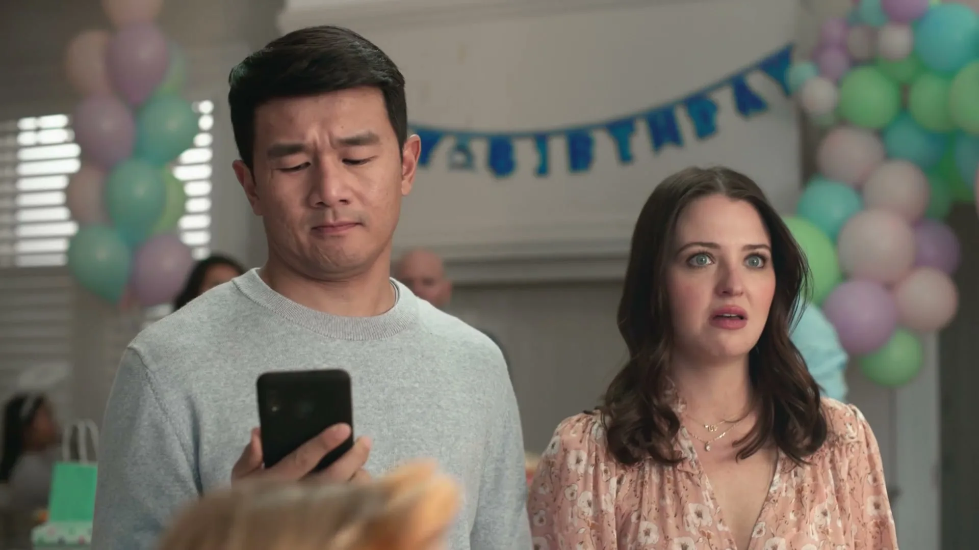 A man and woman stand in a decorated room, the man holding a phone, as the woman reacts to something off-screen.