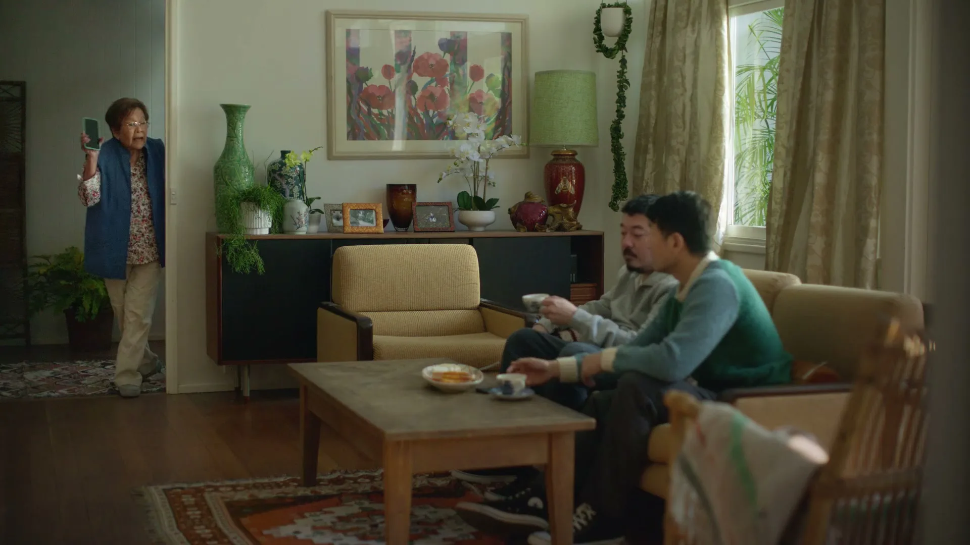 Two men sit on a couch in a living room, looking at a phone, as an older woman enters the room holding a phone.