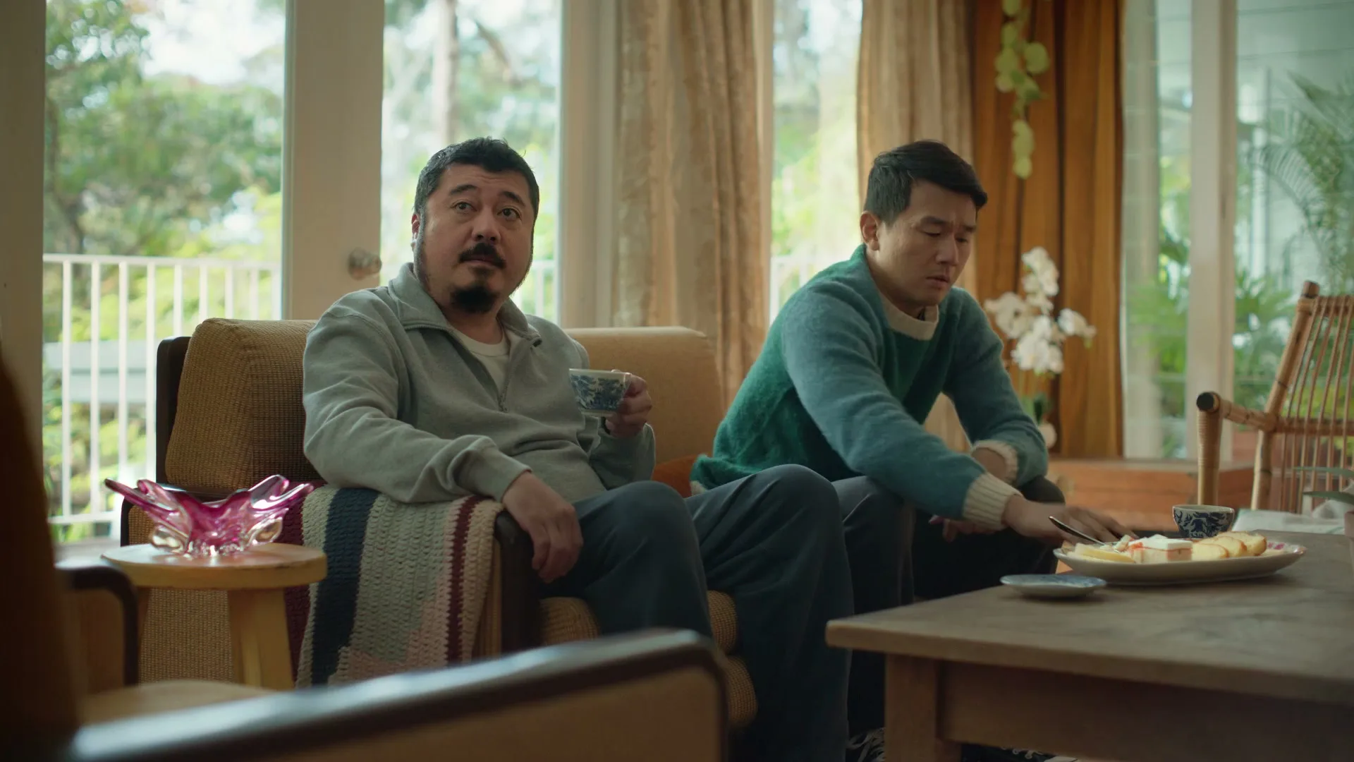 Two Asian men sit in a living room, one on a couch holding a teacup, the other on a chair looking at his phone.