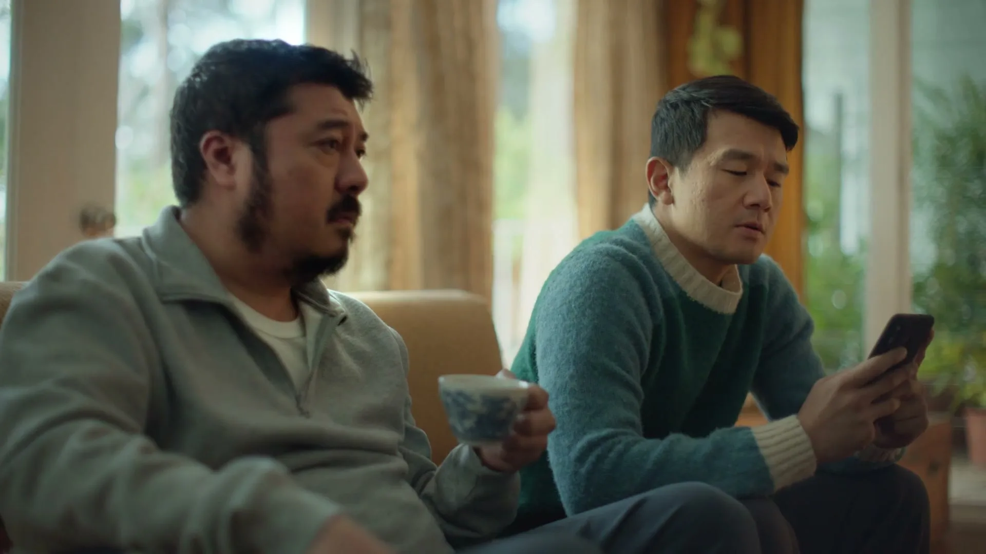 Two men sit on a couch in a living room, one holding a teacup and looking left, the other looking at his phone.