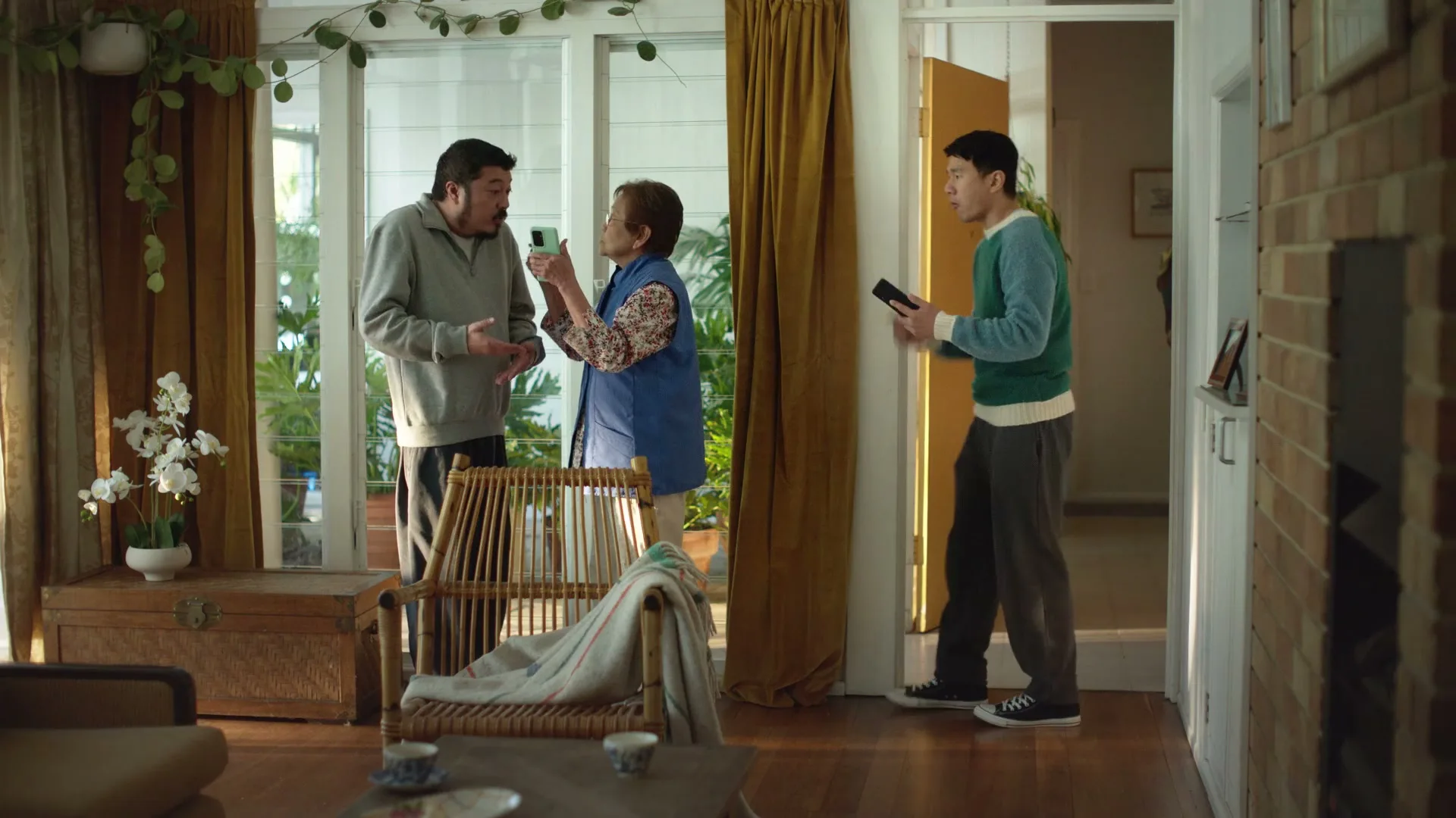 An older man and woman argue over a phone while a younger man looks on in a sunlit living room.