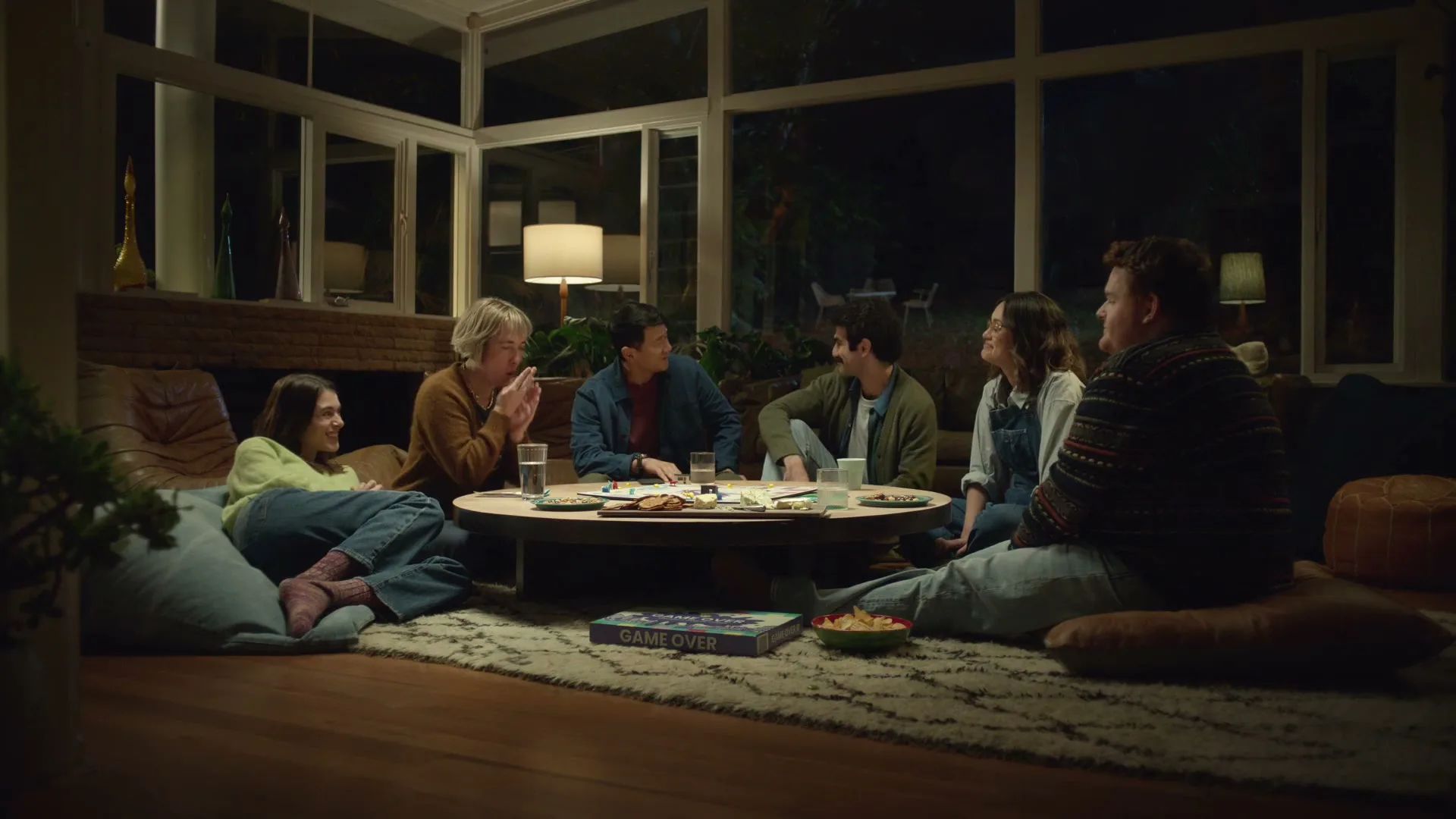 A diverse group of six friends sits on the floor around a coffee table, playing a board game in a modern living room at night.