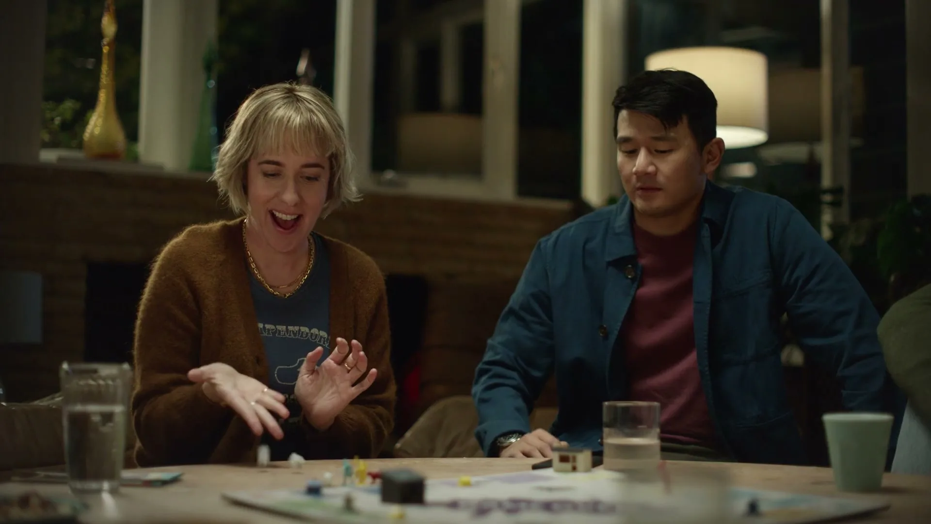 A blonde woman in a brown cardigan claps her hands while a man in a blue jacket looks at his phone at a board game night in a living room.