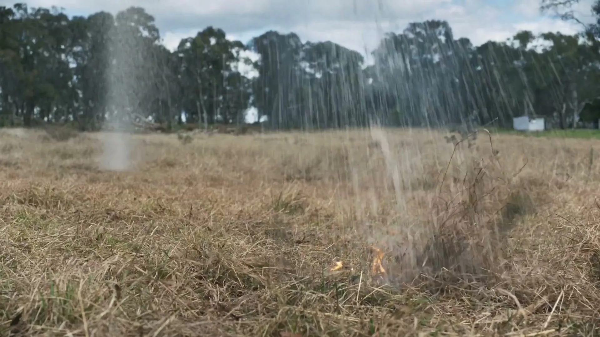 A small fire burns in dry grass in a field, then is extinguished by a stream of water from off-screen.