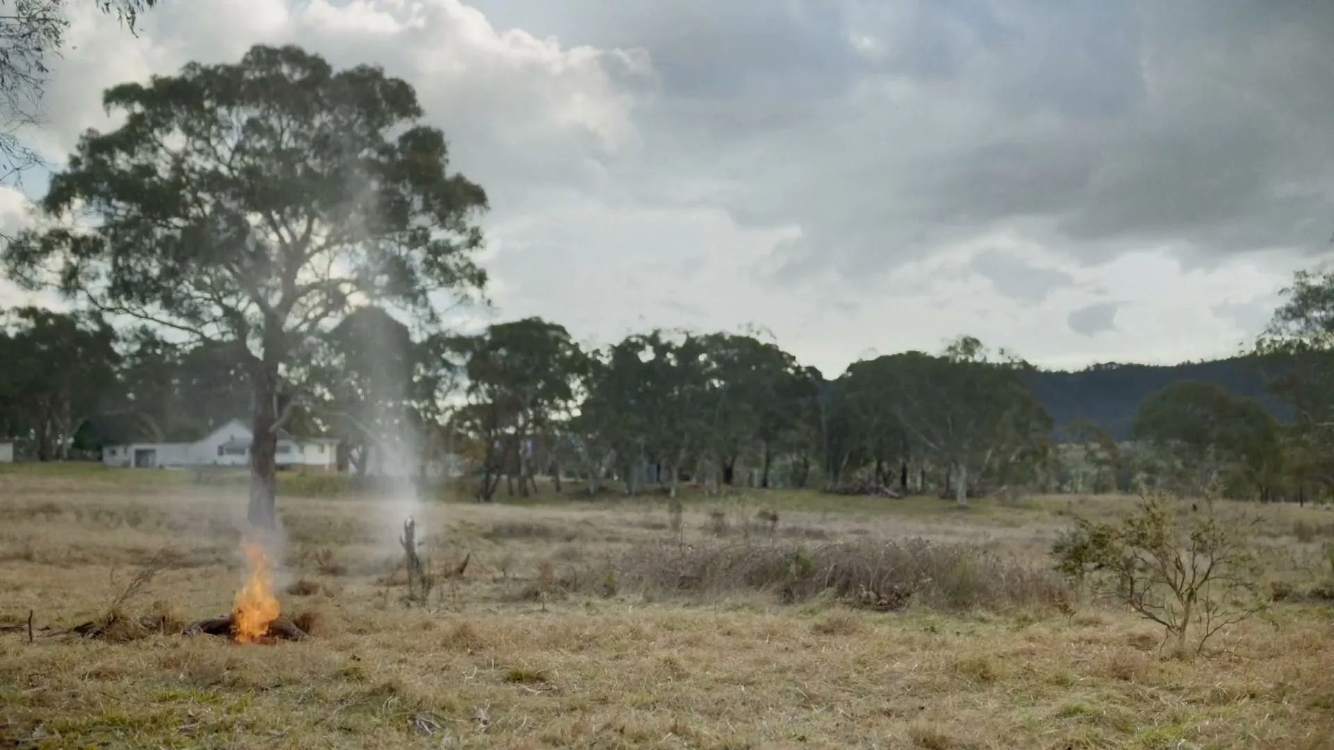 A wide shot of a rural landscape shows a small bonfire burning in a dry field, with a stream of water hitting it from above, under a cloudy sky.
