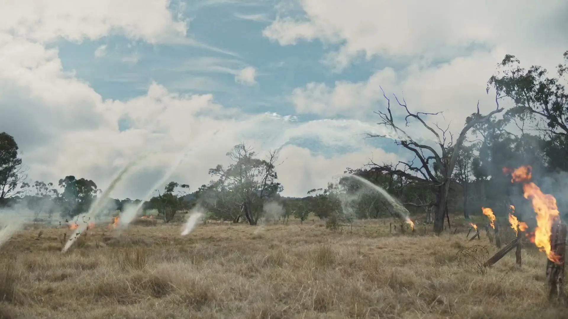 Water streams from multiple sprinklers, fighting small fires in a dry, grassy field under a cloudy sky.