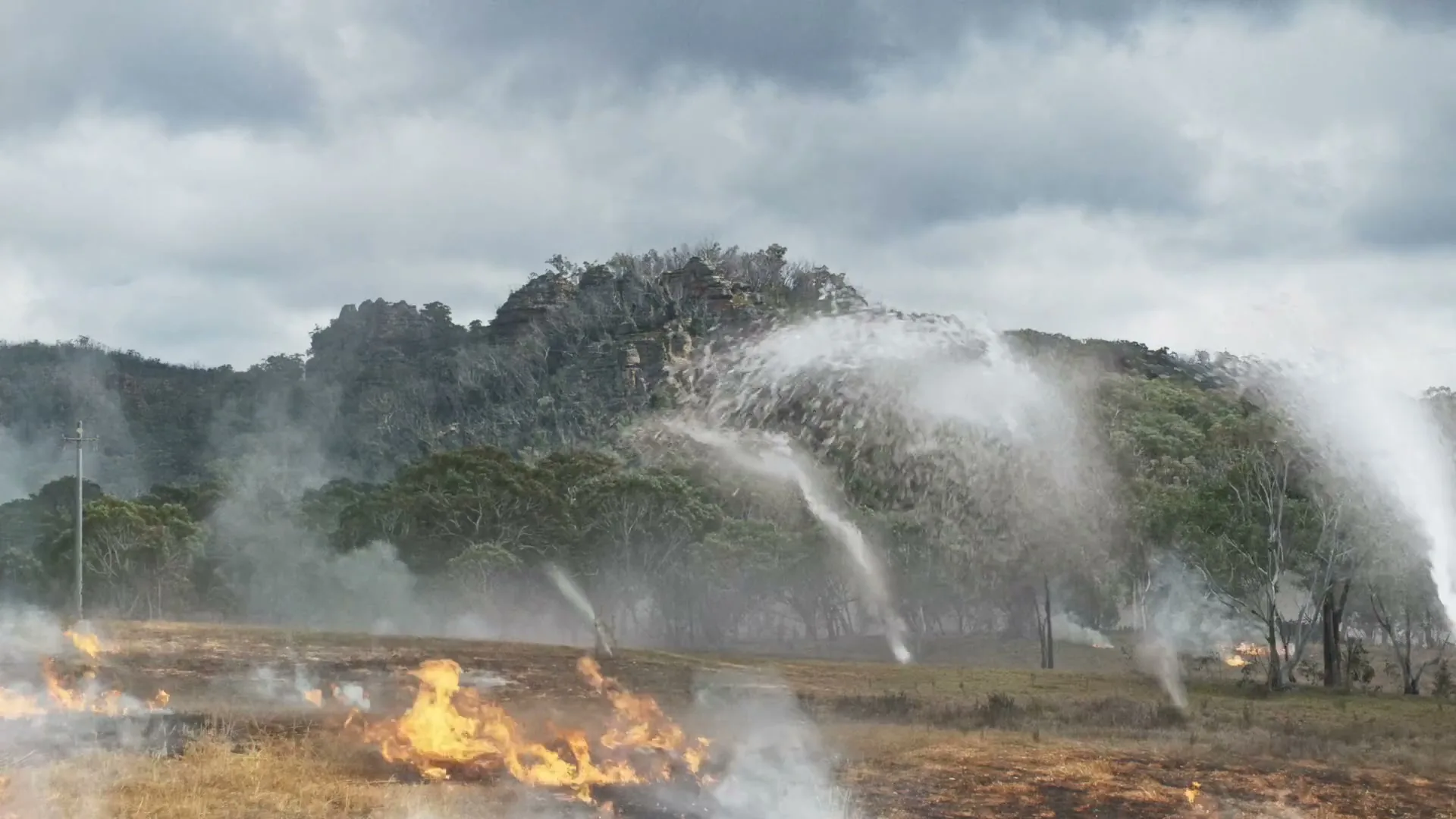 A wide shot shows a dry field with patches of fire and smoke, while multiple water streams spray across the landscape, with a rocky, tree-covered hill in the background under a cloudy sky.
