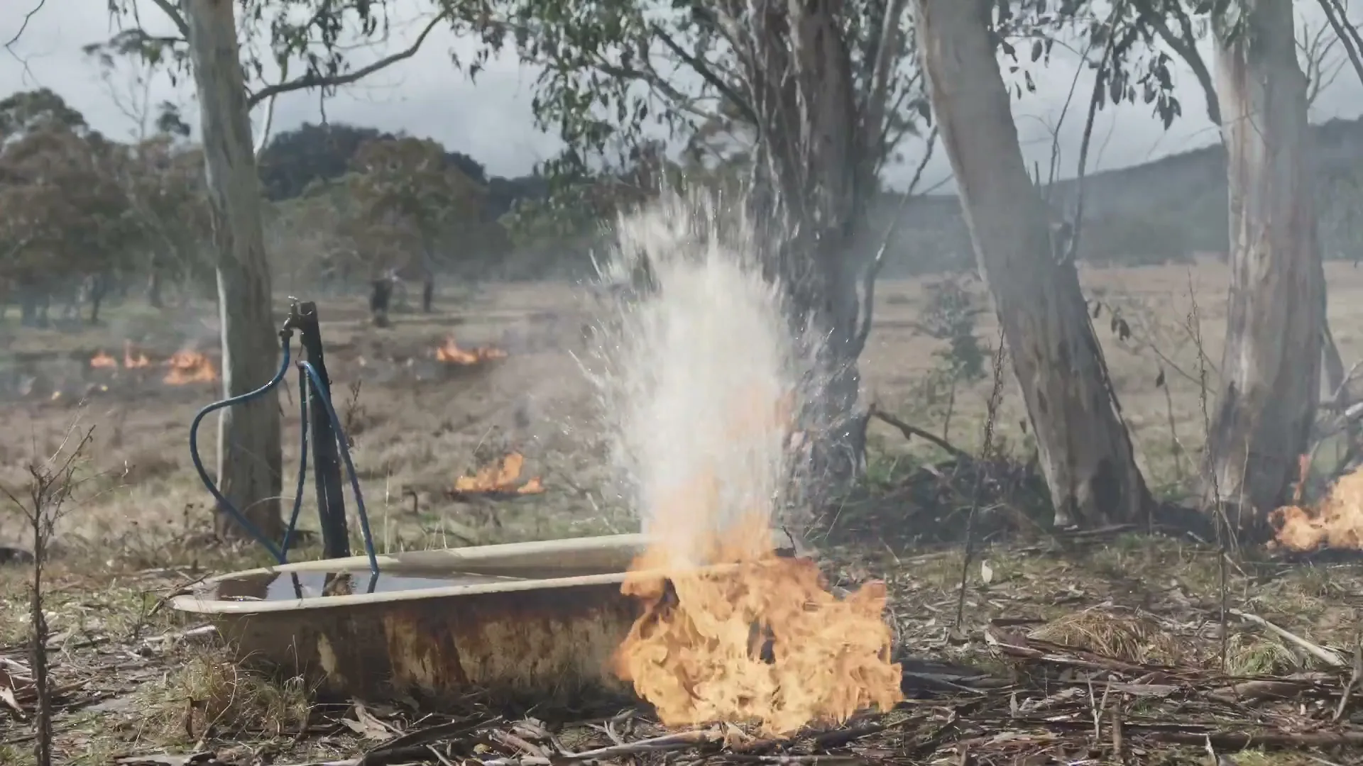 A metal trough with water and fire around it, as water sprays upwards from a blue hose in a dry, smoky field with trees and hills.