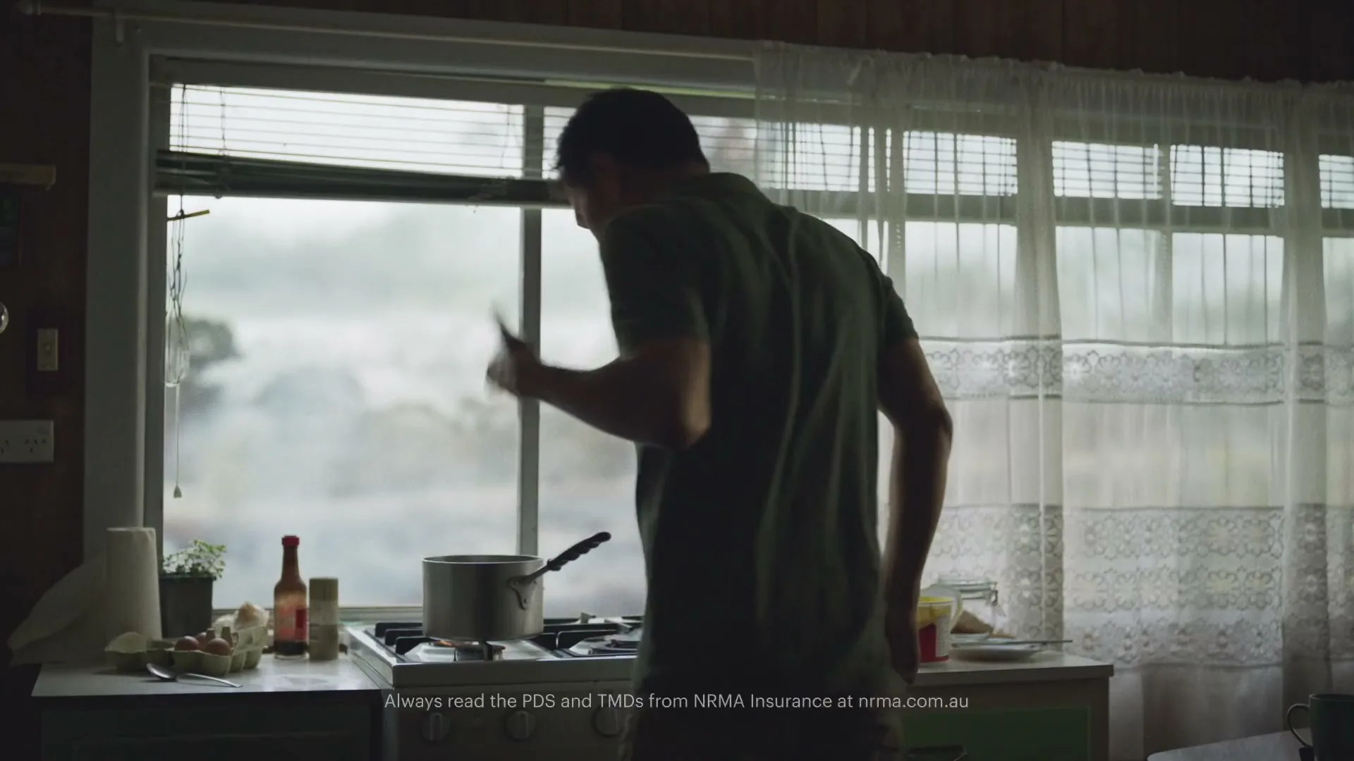 A man in a green shirt stands with his back to the camera, stirring a pot on a stove in a rustic kitchen, with a window showing a hazy exterior.