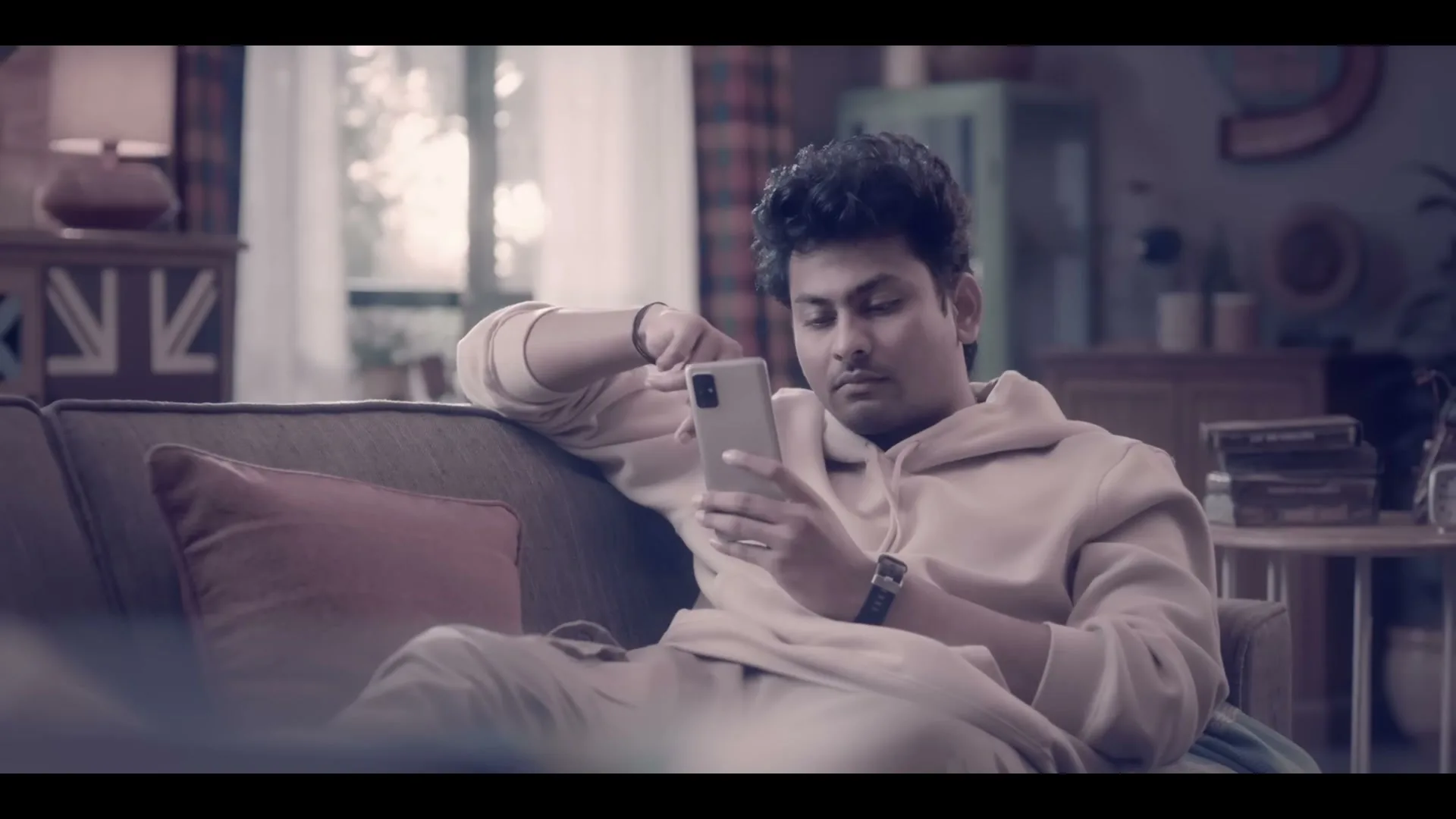 A young man with curly hair, wearing a beige hoodie, sits on a sofa and looks at his phone in a living room.