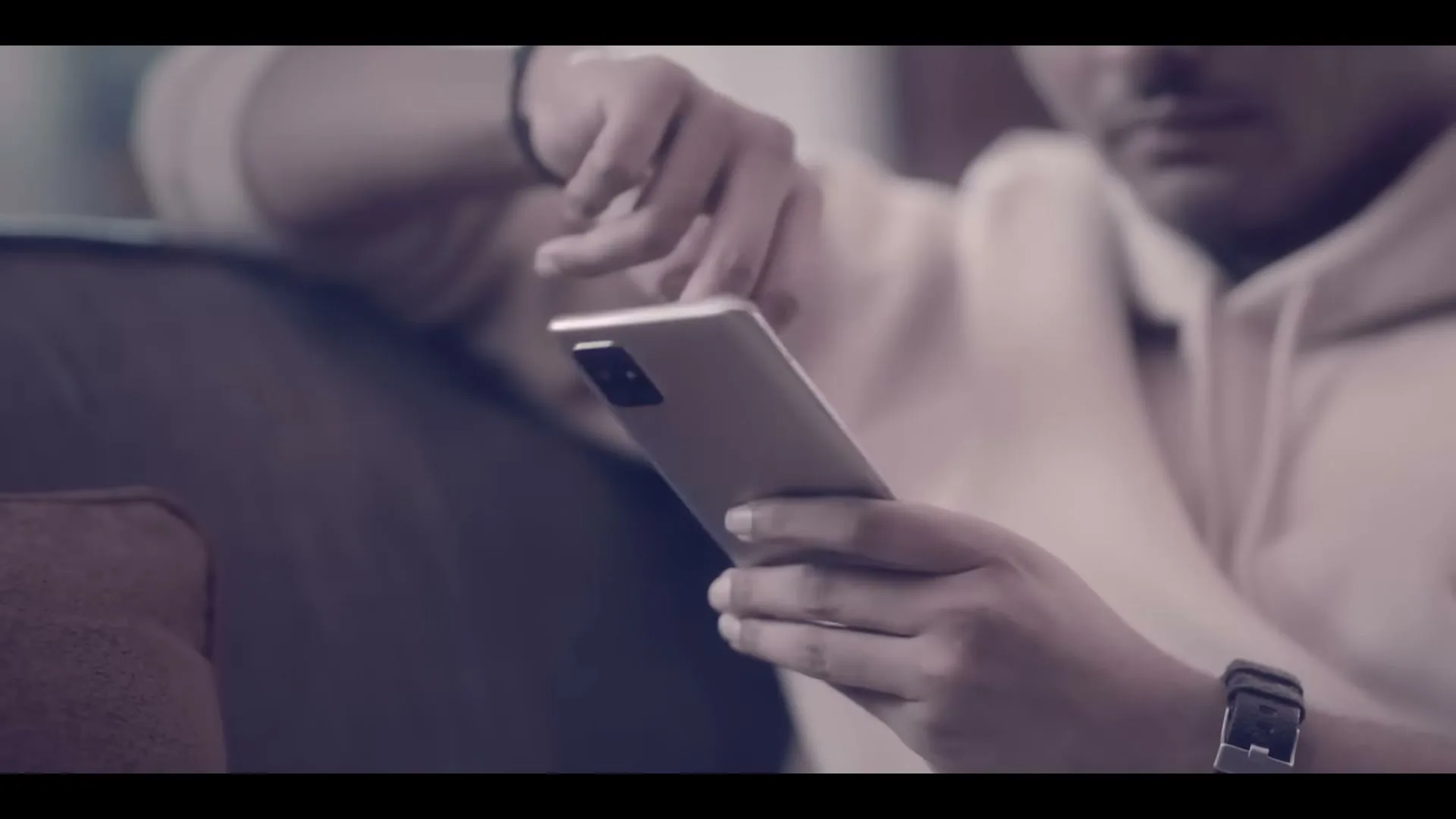 A young man in a hoodie sits on a couch, looking at his phone in a dimly lit interior.