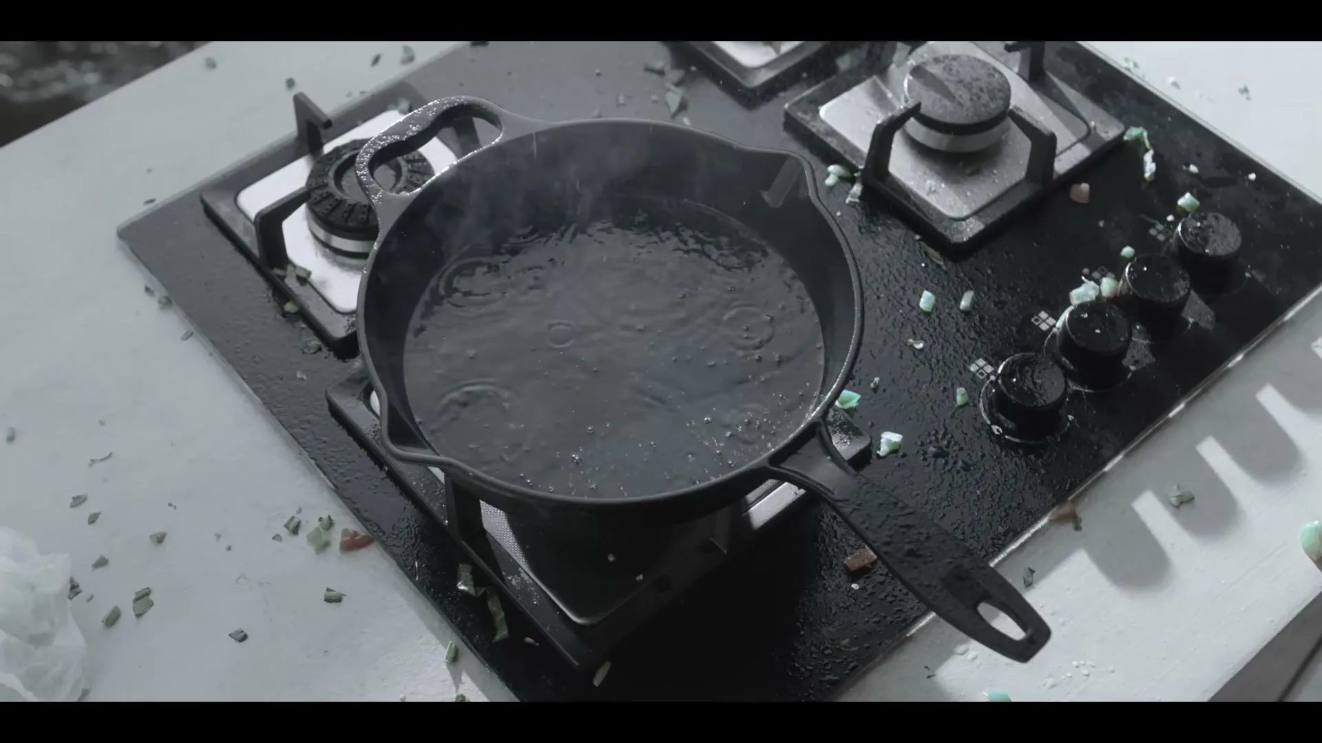 A black cast iron pan with water boiling in it sits on a black stovetop, surrounded by small, colorful debris in a kitchen.
