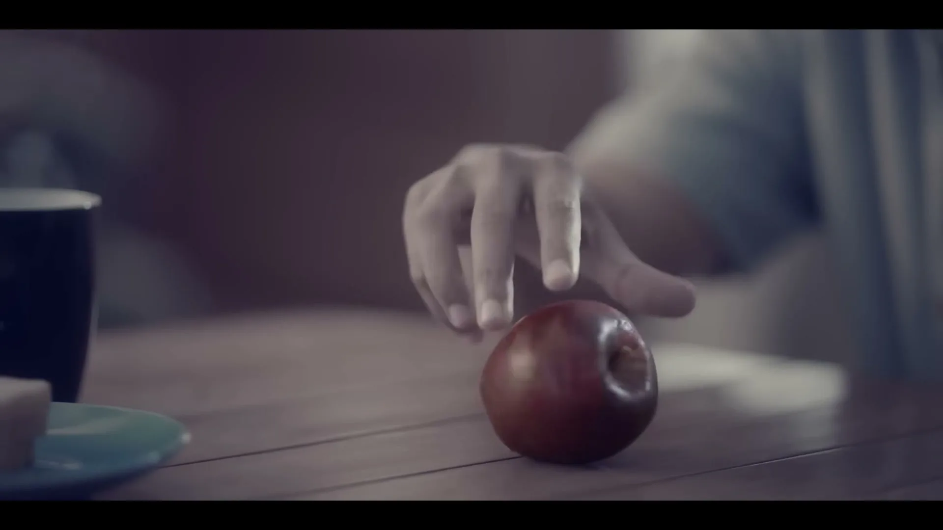 A person's hand reaches for a red apple on a wooden table, with a mug and plate nearby, in a dimly lit interior.