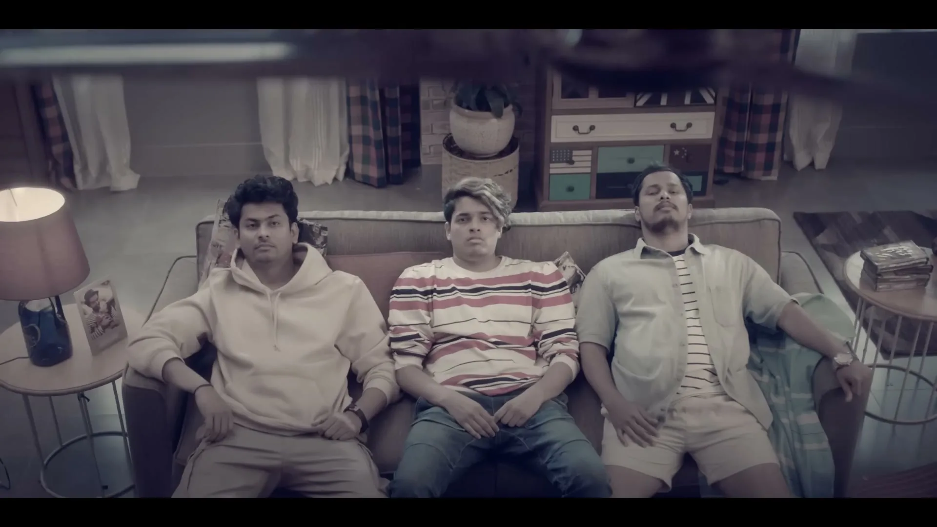 Three young men sit on a couch, looking tired and disheveled, as a blurred object passes in front of them in a living room.