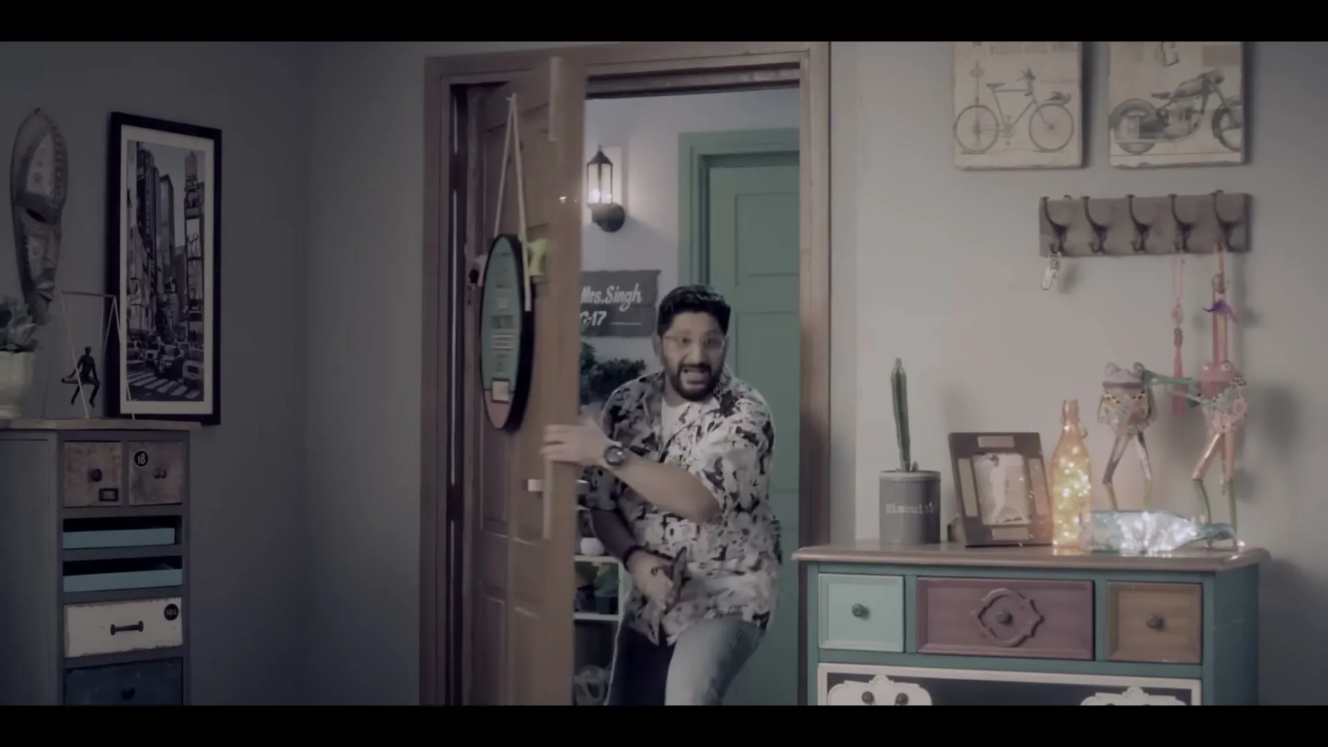 A man in a patterned shirt and jeans bursts through a wooden door into a room with decorative furniture and wall art.