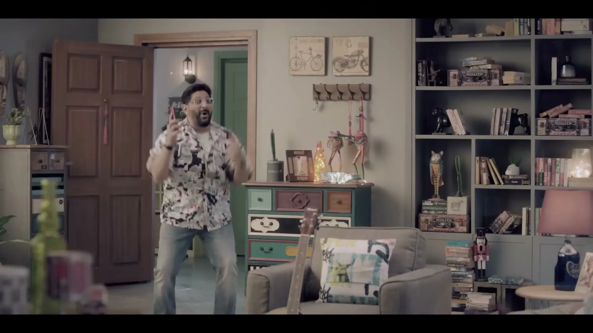 A man in a patterned shirt and jeans stands in a living room, gesturing excitedly with his hands while talking on the phone.