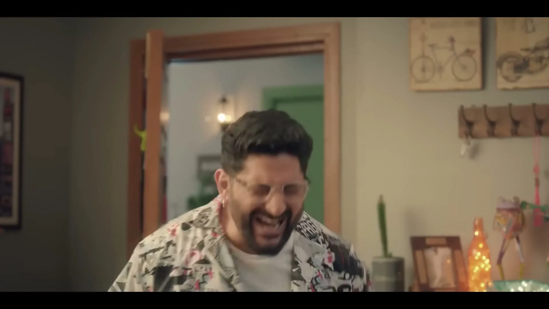 A man with a beard and glasses, wearing a patterned shirt, laughs excitedly in a living room.