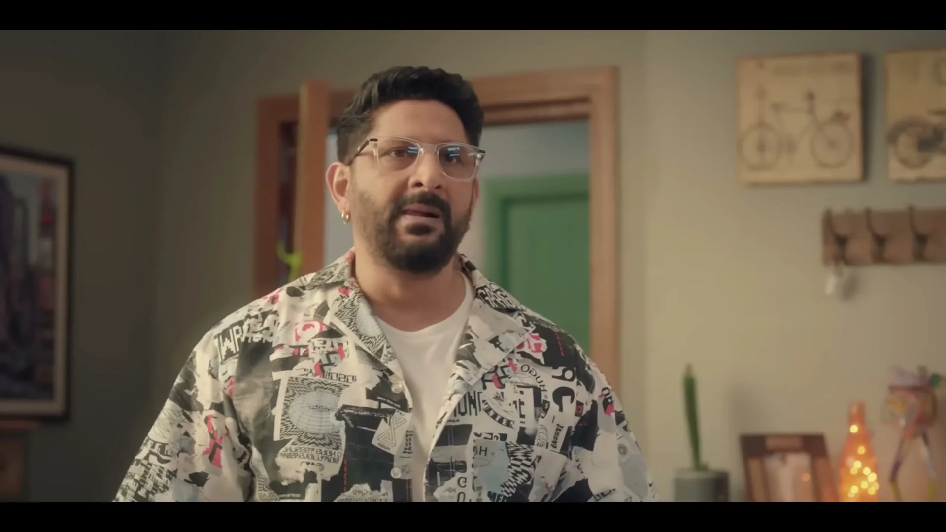 A man with a beard and glasses, wearing a patterned shirt, stands in a room and speaks, looking directly at the camera.