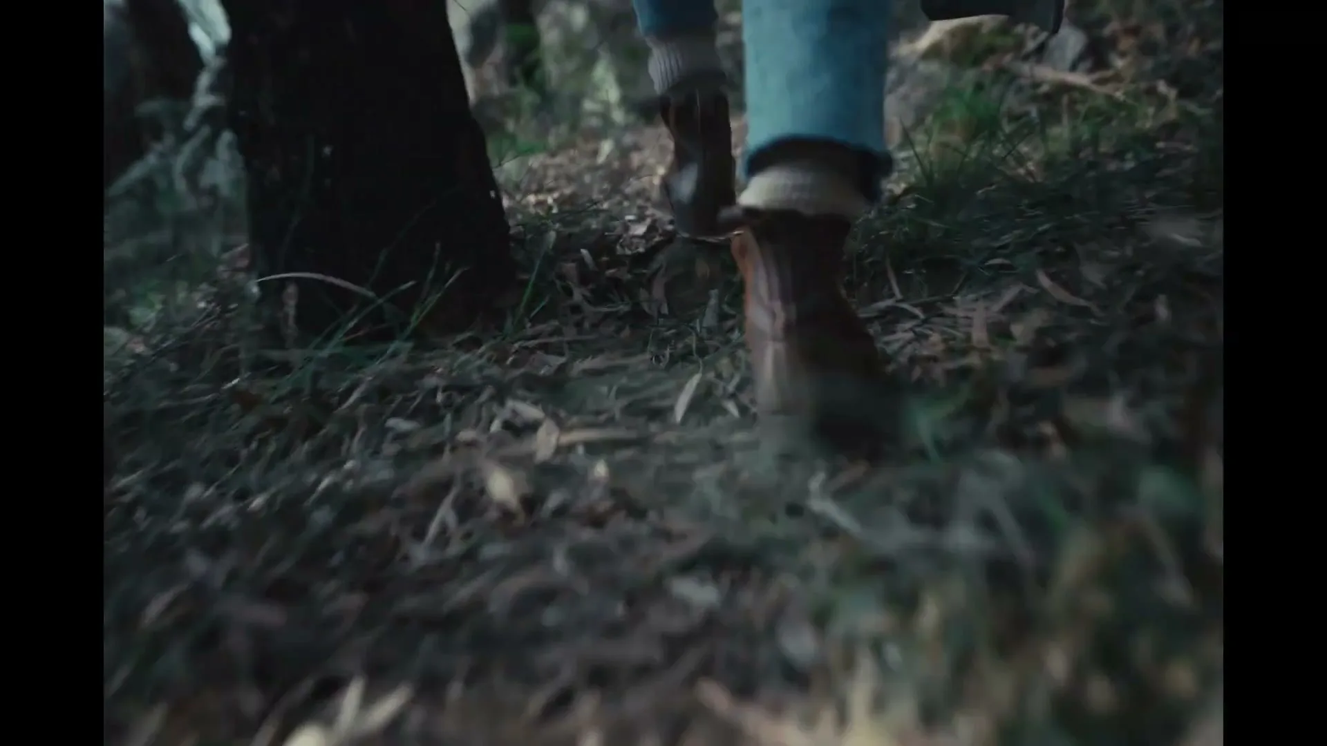 A woman in jeans and boots walks through a forest, carrying an axe over her shoulder, with rocks and trees in the background.