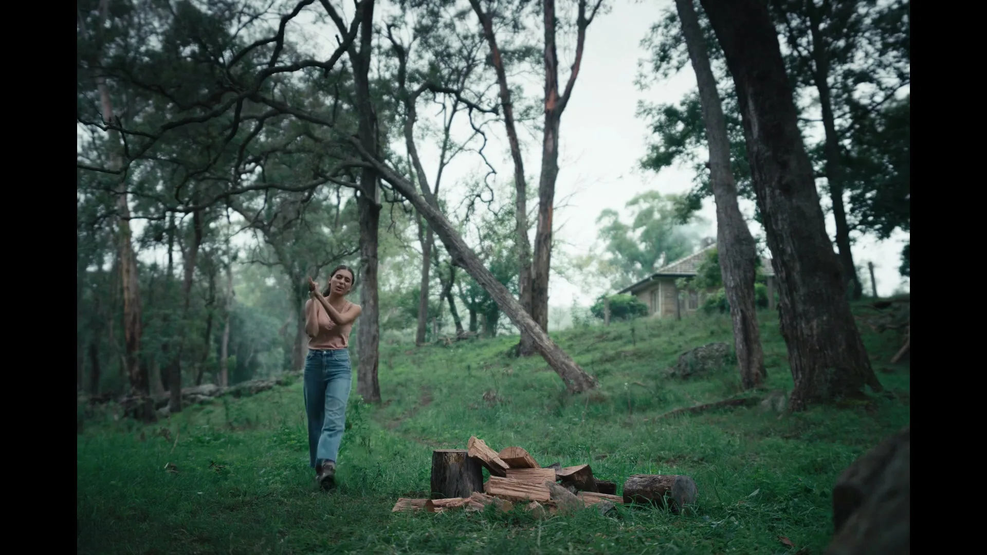 A young woman in a tank top and jeans chops wood with an axe in a lush, green forest with a small house visible in the background.