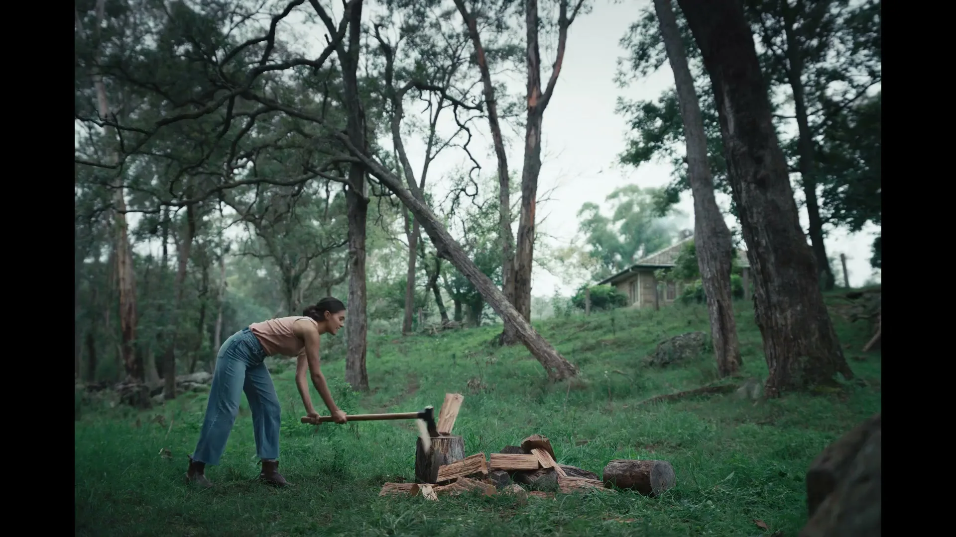 A young woman in a tank top and jeans chops wood with an axe in a forest clearing, with a rustic house visible on a hill in the background.