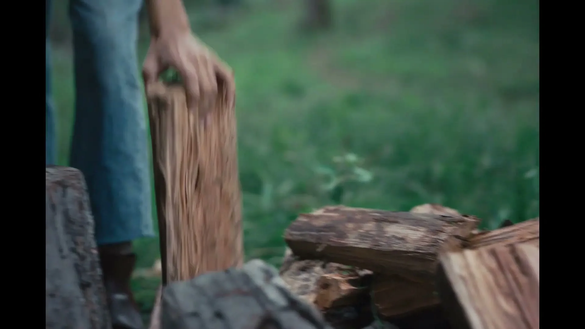 A person in blue jeans picks up a piece of firewood from a pile and places it on a tree stump in a grassy outdoor area.