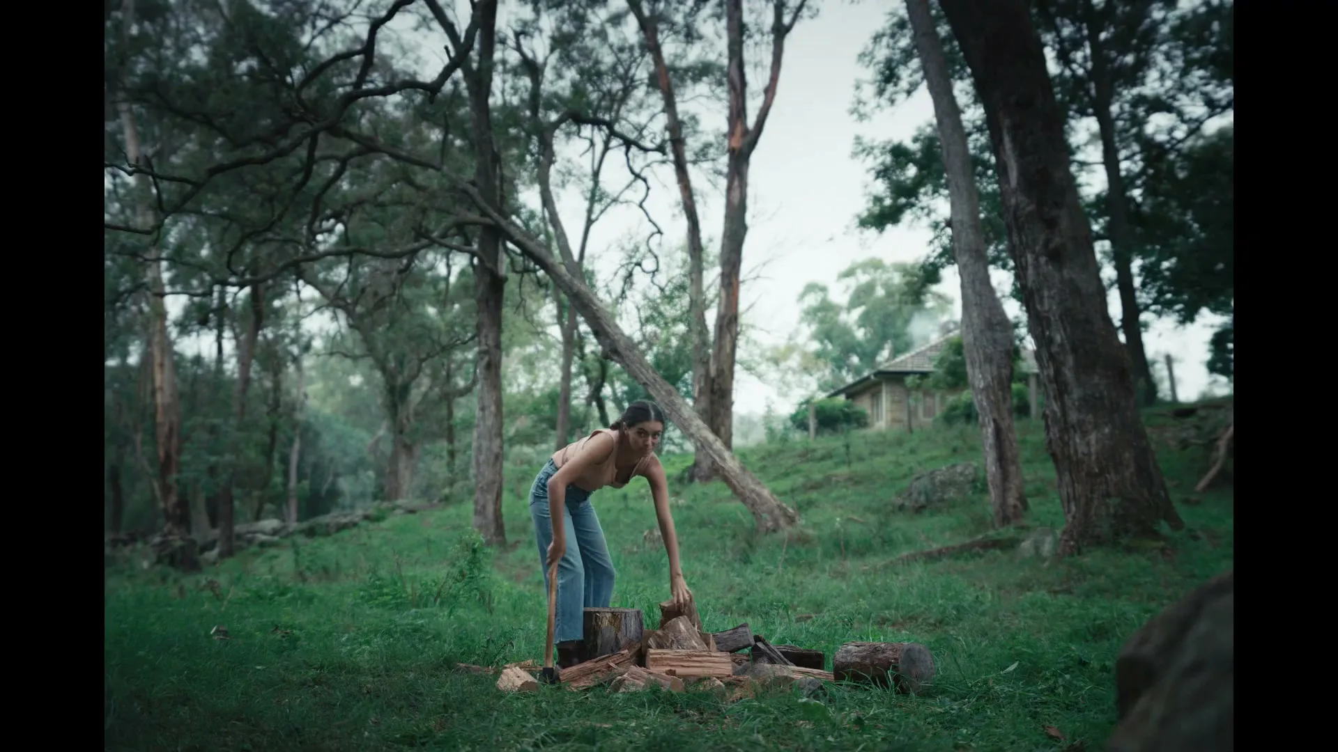 A young woman in a tank top and jeans chops firewood with an axe in a grassy, wooded area with a small house in the background.